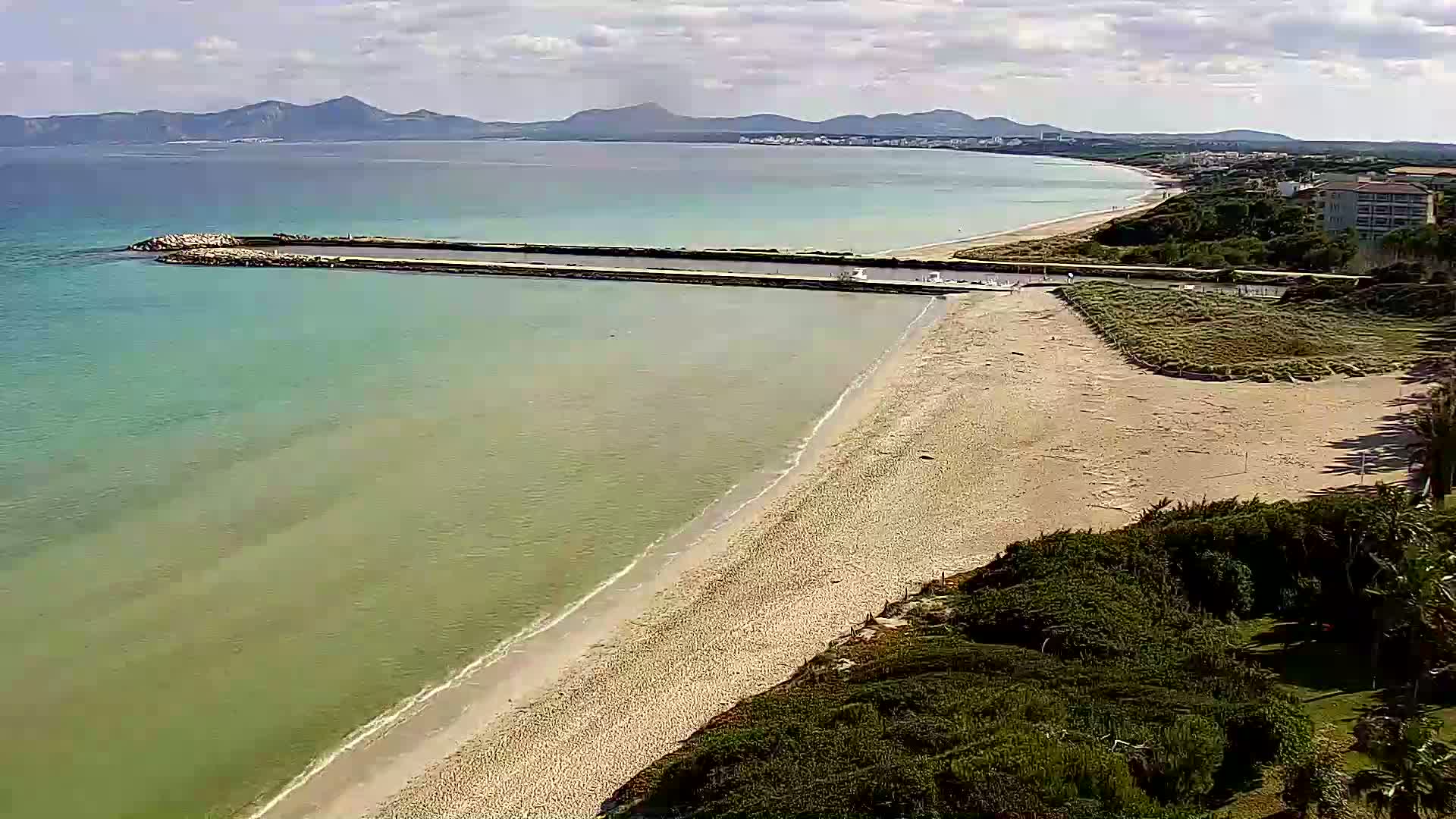 An aerial view shows a sandy beach meeting a calm, light-green sea, a man-made breakwater, and distant mountains under a partly cloudy sky.