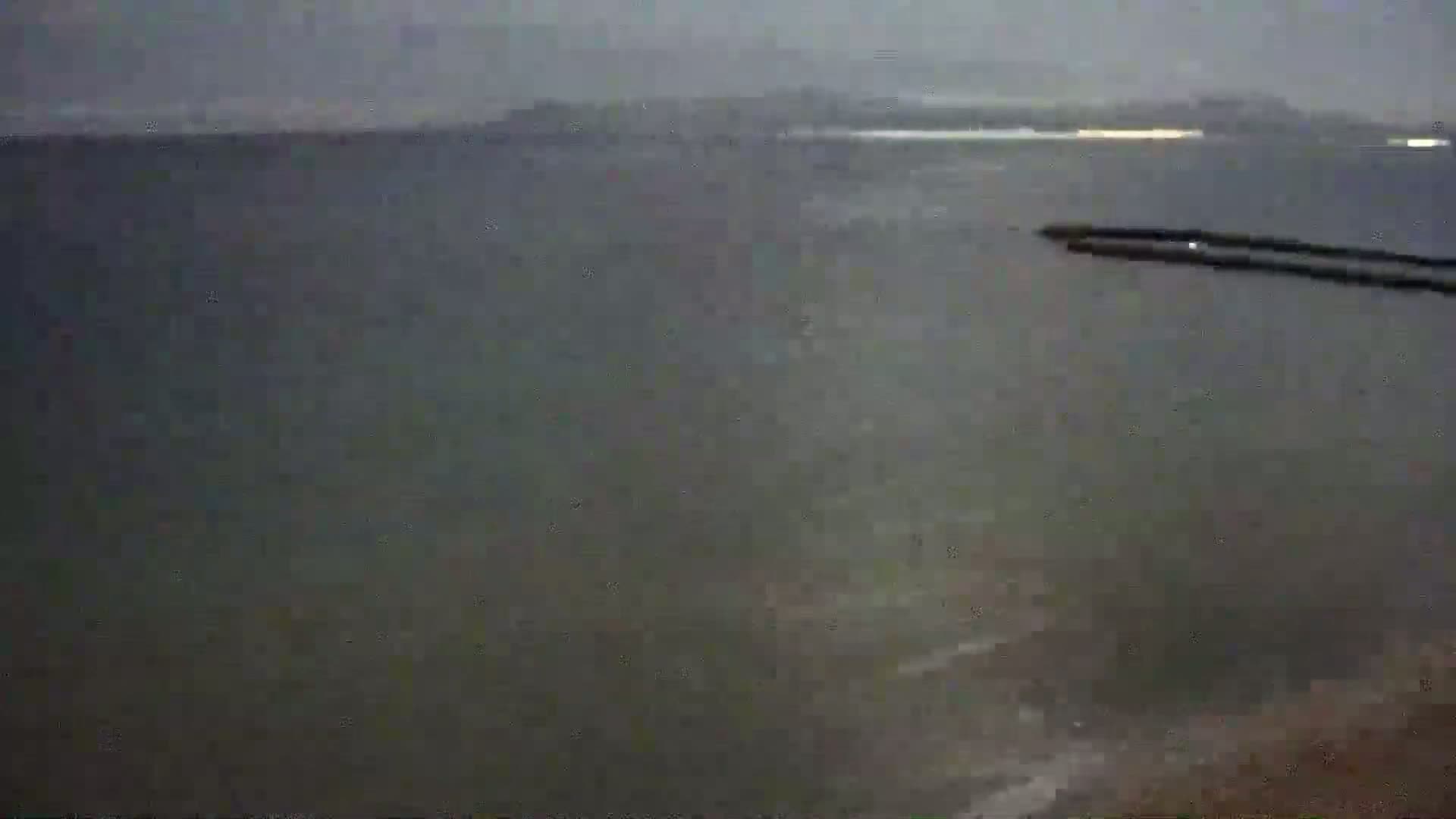 A dark, overcast view of a body of water at night, with a breakwater visible in the distance and faint lights on the far shore.