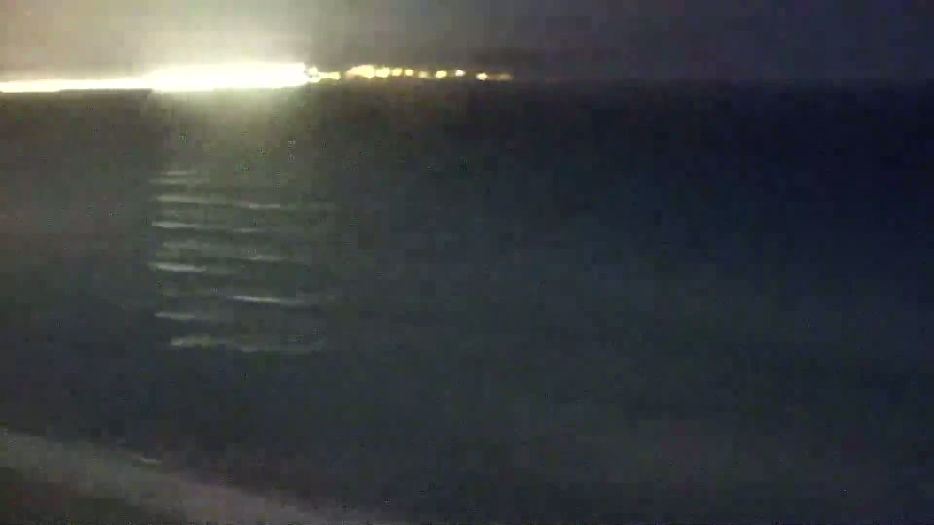 A nighttime view of dark water with faint waves, illuminated by a distant line of lights on the horizon.