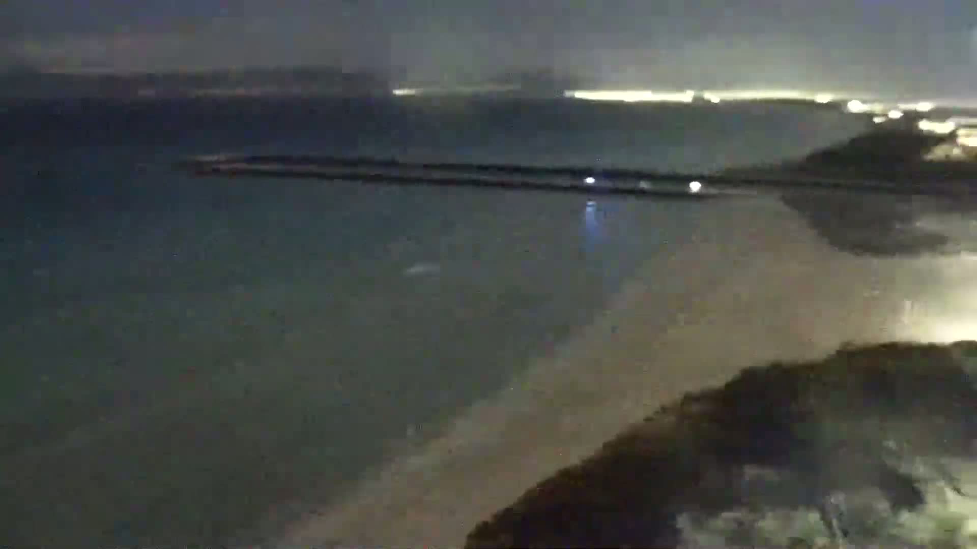 A nighttime view shows a dark body of water with a long, low structure extending from the shore, a sandy beach, and dark land in the foreground.