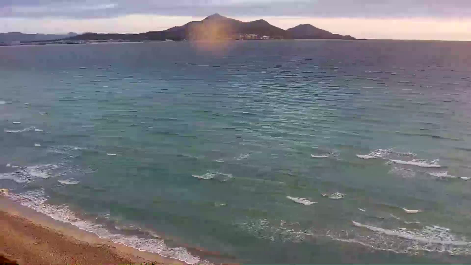A sandy beach with gentle waves laps against the shore, under a partly cloudy sky, with a distant mountain range visible across the calm sea.