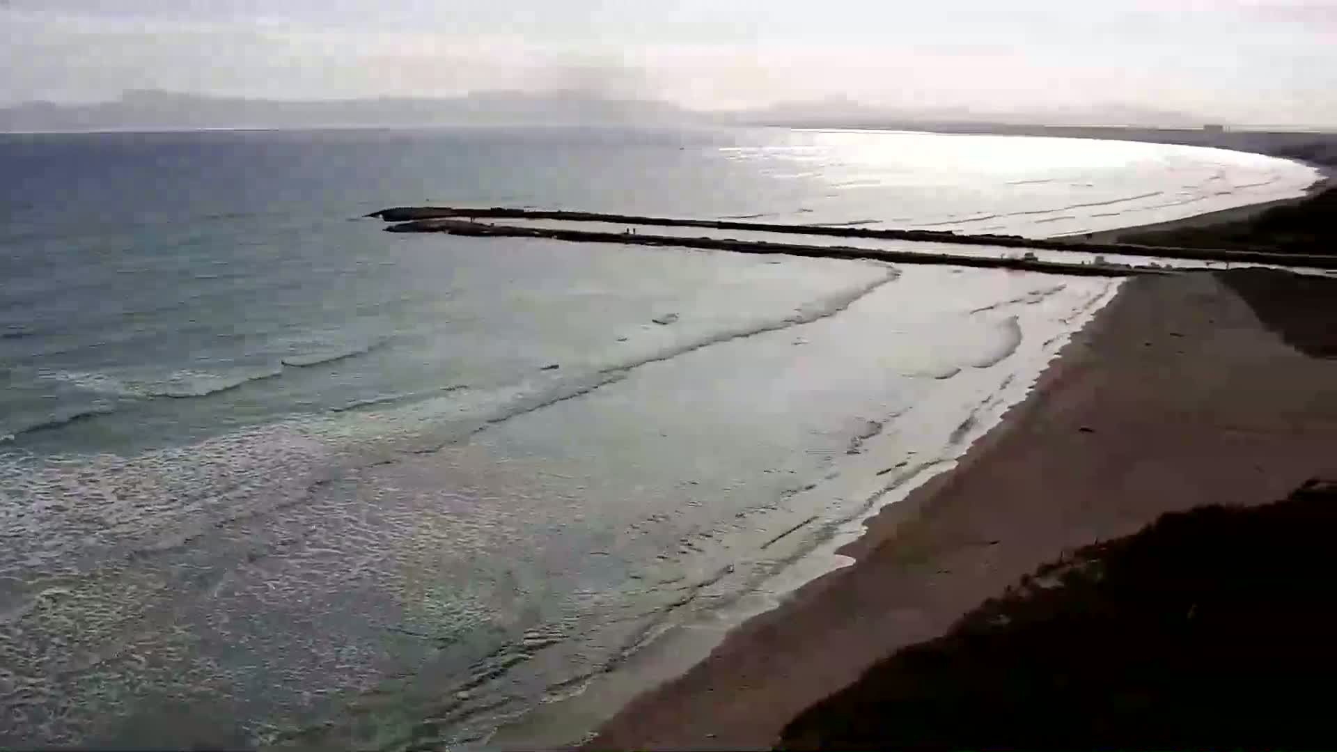 A long sandy beach curves gently around a calm sea, where a man-made jetty extends into the water under a mostly cloudy sky.