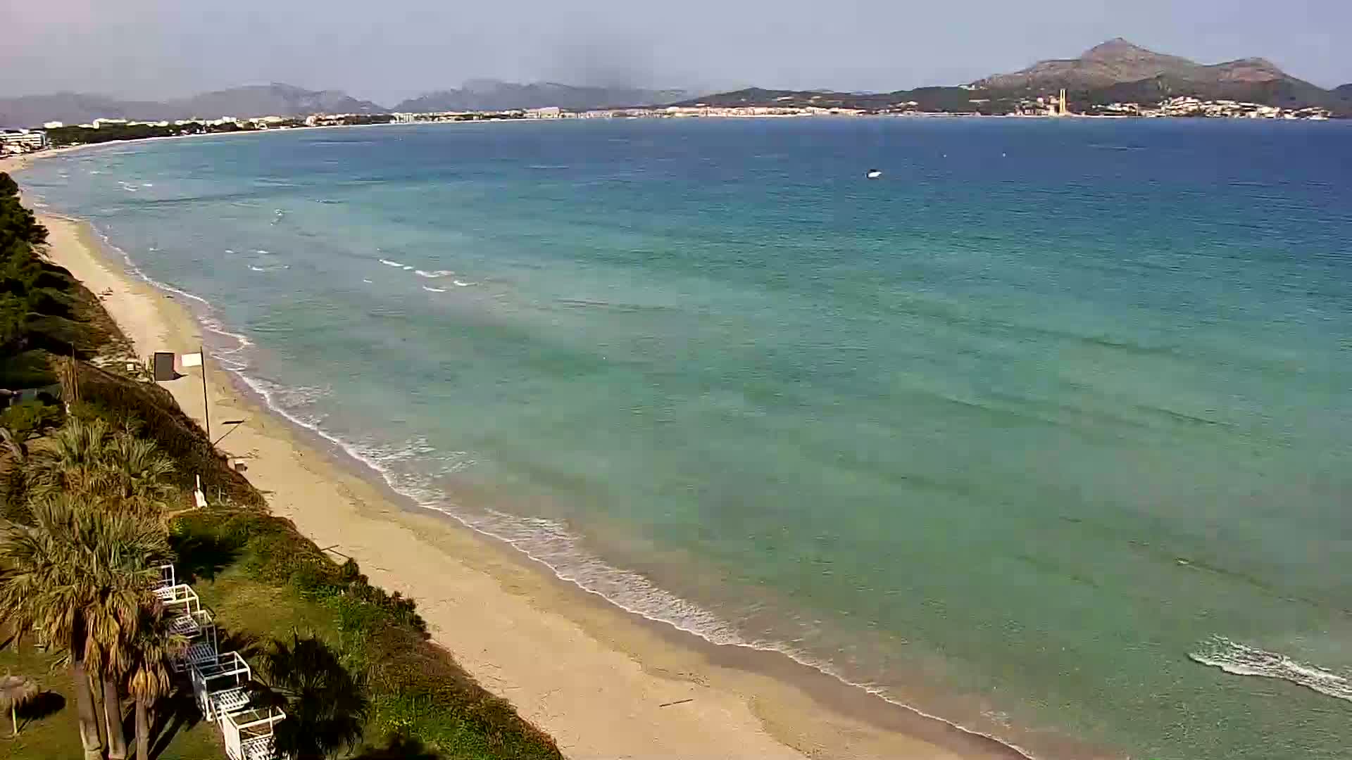A sandy beach with gentle waves laps a turquoise sea under a mostly clear sky, with distant mountains visible in the background.