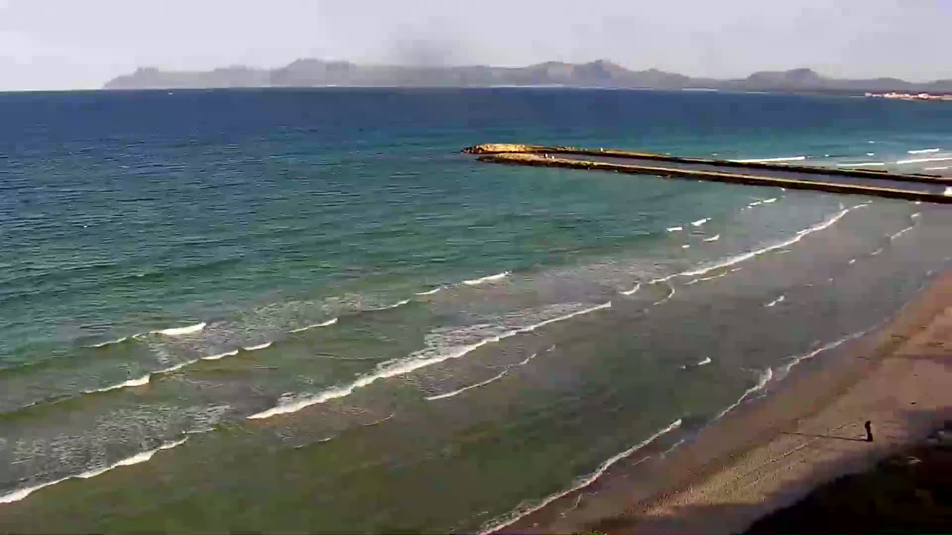 A sandy beach with gentle waves rolls onto the shore, a long jetty extends into a calm sea under a mostly clear sky with distant mountains visible on the horizon.