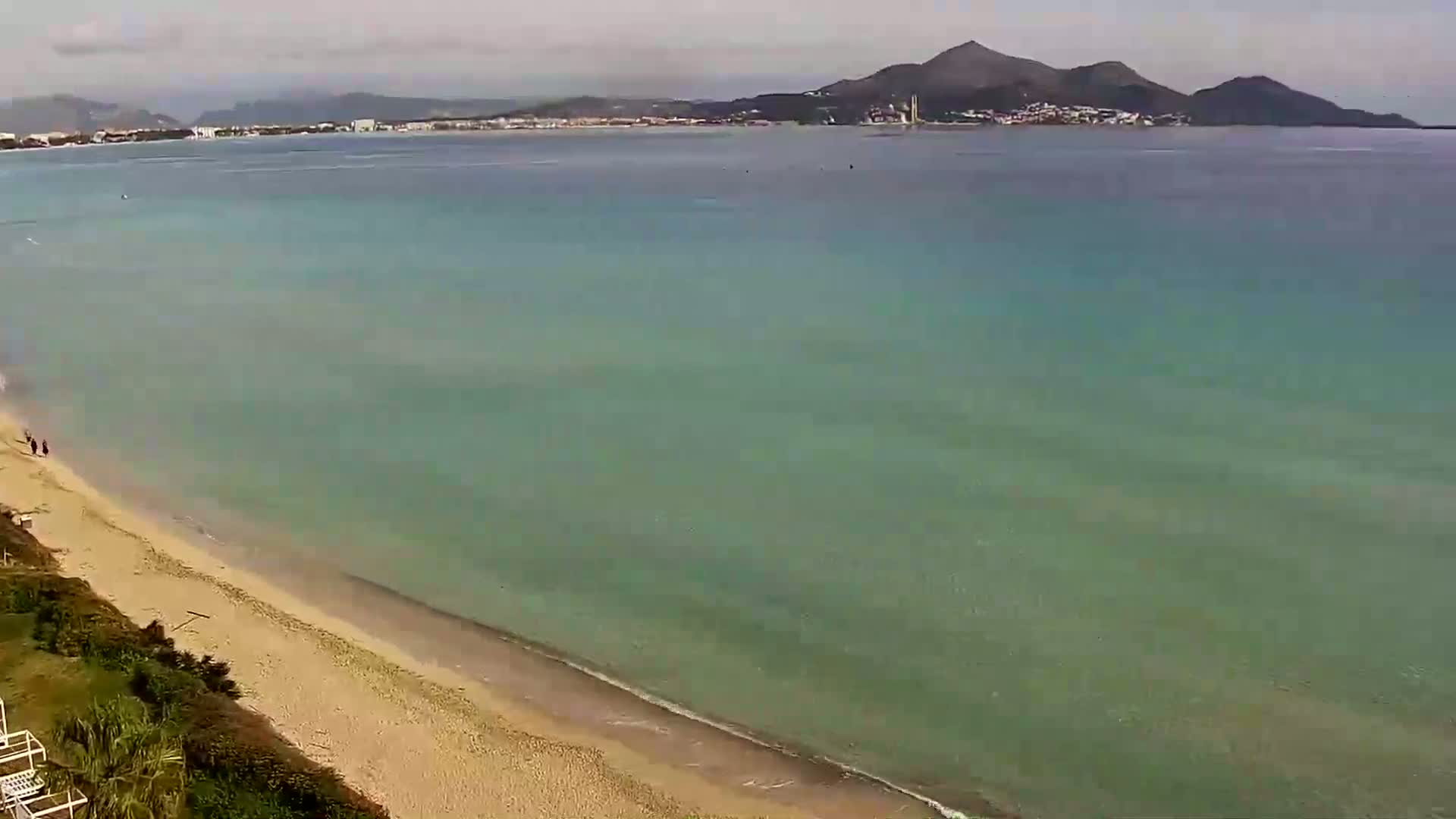 A sandy beach slopes gently into a calm, clear teal sea, with a distant mountain range visible under a cloudy sky.