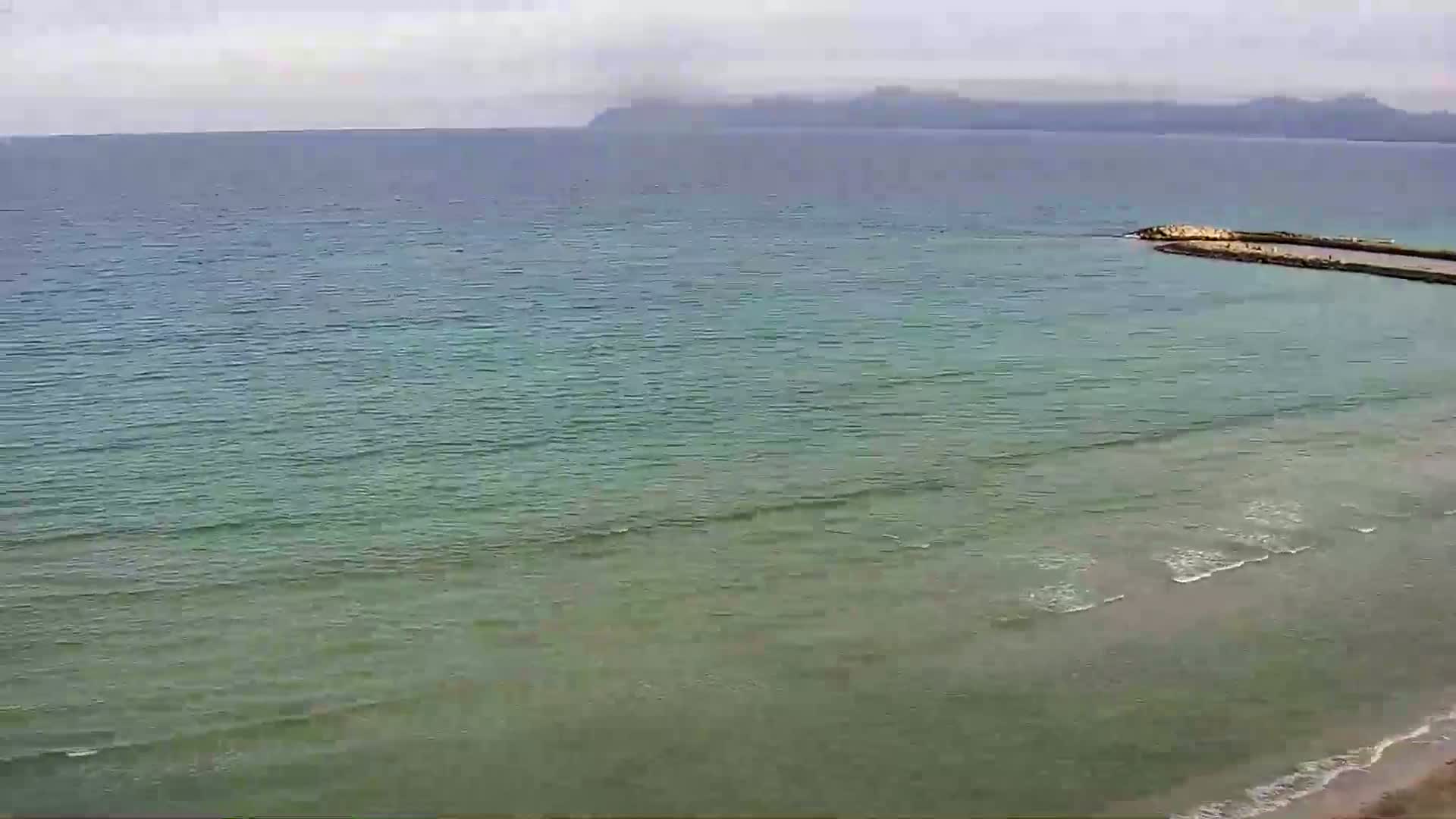 A calm, light-green sea meets a sandy beach under an overcast sky, with a small man-made jetty extending into the water and distant mountains visible on the horizon.