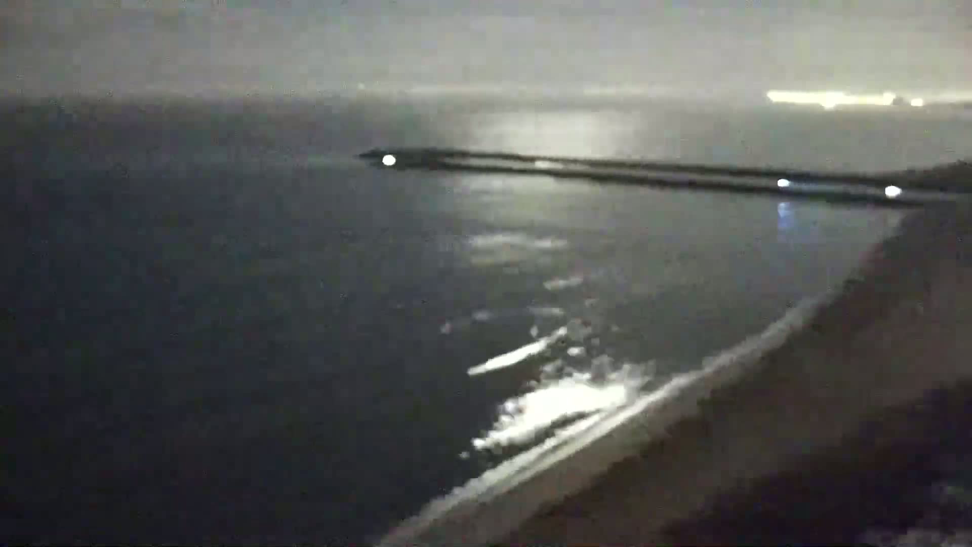 A dark, nighttime coastal scene shows a long, dark breakwater extending into a somewhat choppy sea under an overcast sky.