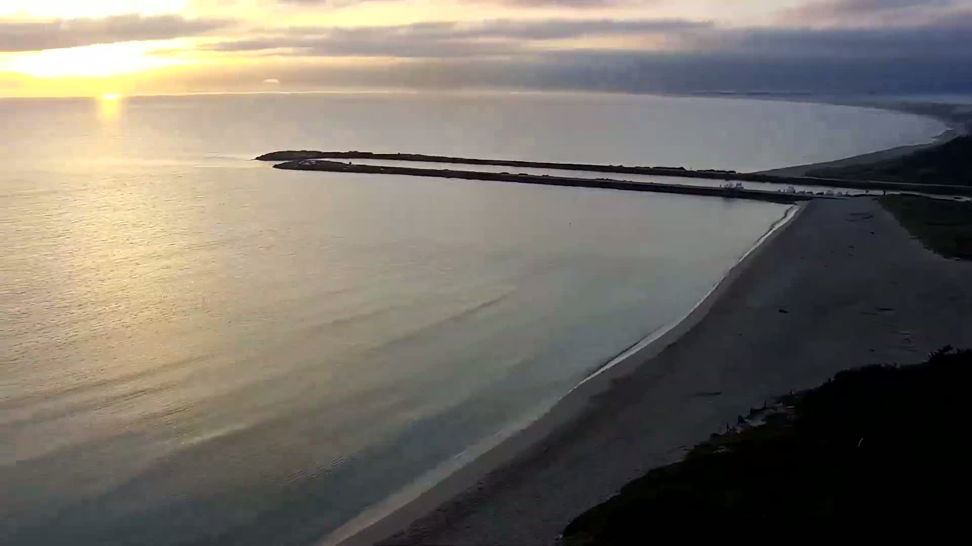 A calm ocean at sunrise meets a sandy beach bordered by dark vegetation and a man-made breakwater.