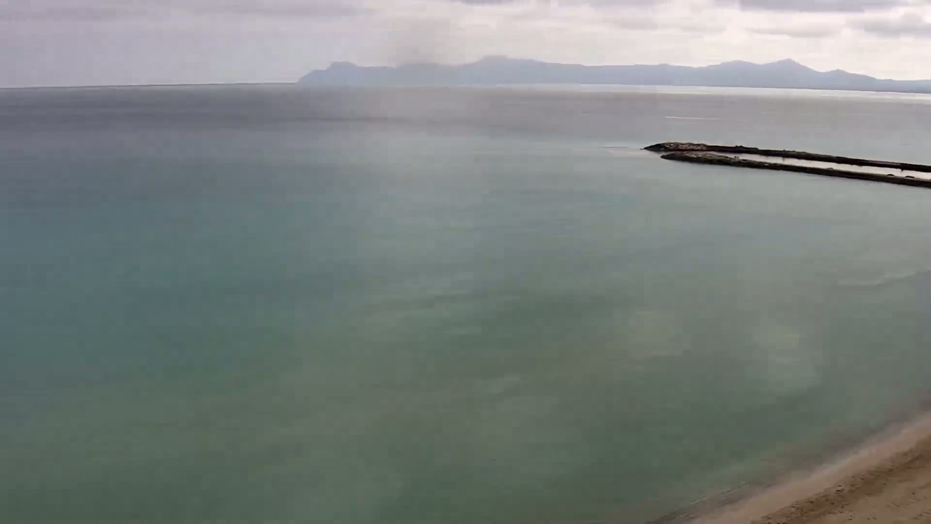 A calm, light-green sea stretches to a distant mountain range under a cloudy sky, with a small jetty visible in the middle distance and a sandy beach in the bottom right corner.
