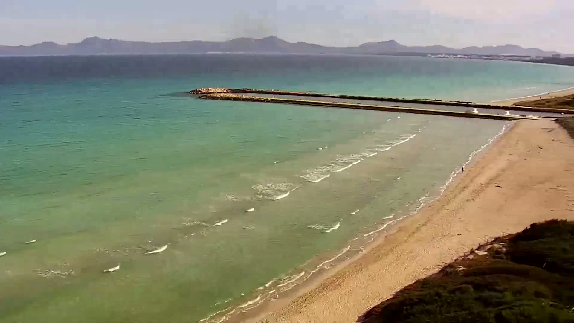 An aerial view shows a sandy beach meeting a calm, teal-colored sea on a sunny day, with a long, narrow jetty extending from the shore into the water.