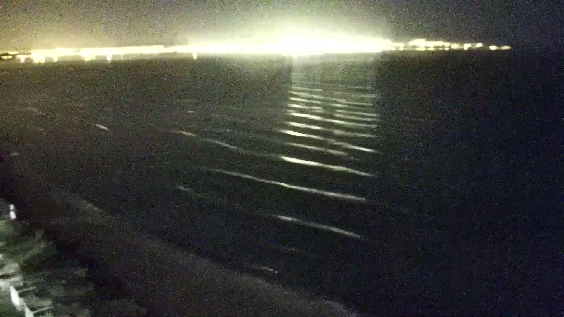 A nighttime view of a dark body of water with small waves reflecting light from a distant, brightly lit shoreline.