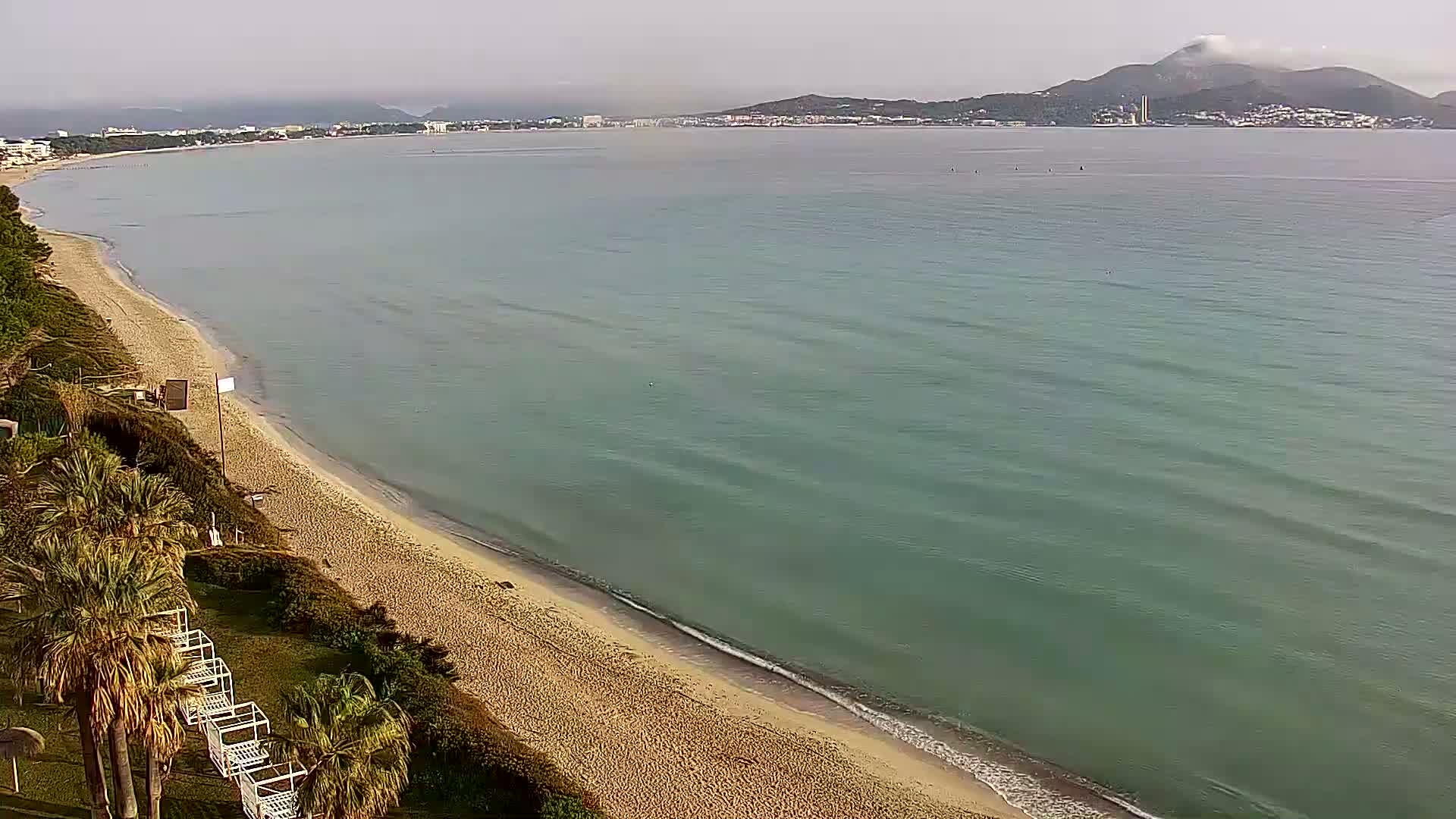 A sandy beach slopes gently into a calm, teal-colored sea under a hazy sky, with a distant mountain range visible in the background.