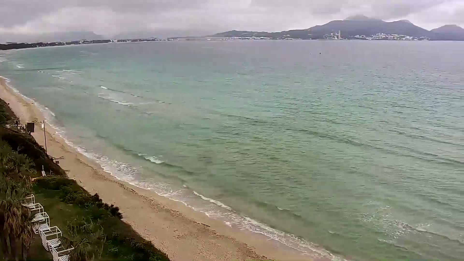 A sandy beach with gentle waves under an overcast sky stretches towards a calm sea and distant mountains.