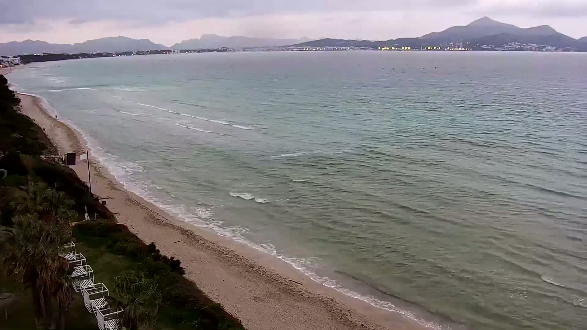 A sandy beach with gentle waves laps the shore under an overcast sky, with mountains visible in the distance.