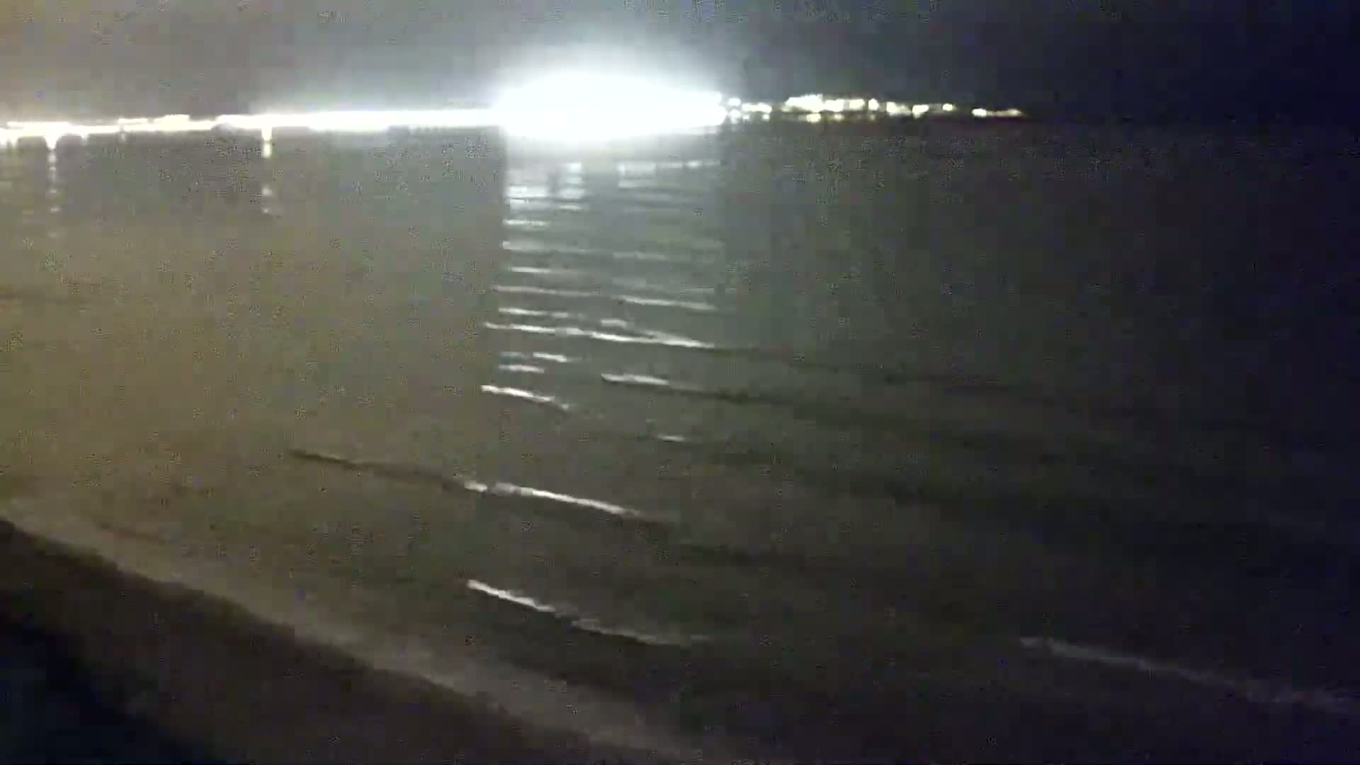 A dark, nighttime beach scene shows gently rolling waves lapping at the shore under a bright, distant light source.