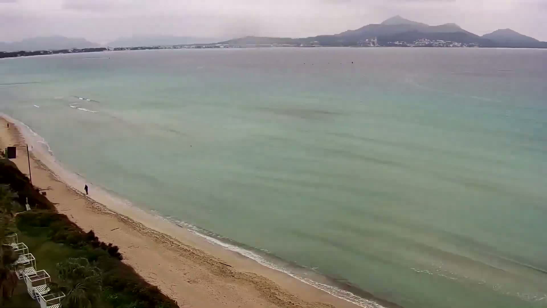 A person walks along a sandy beach next to calm, light-green water under an overcast sky, with mountains visible in the distance.