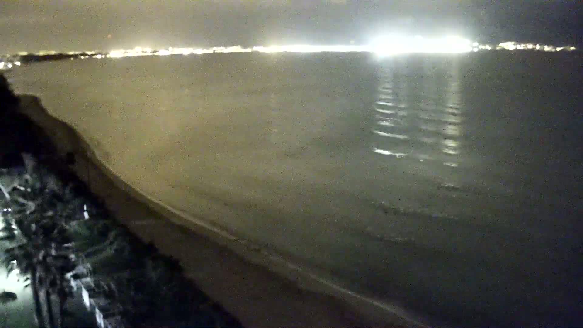 A nighttime view of a calm sea with a sandy beach and distant city lights.