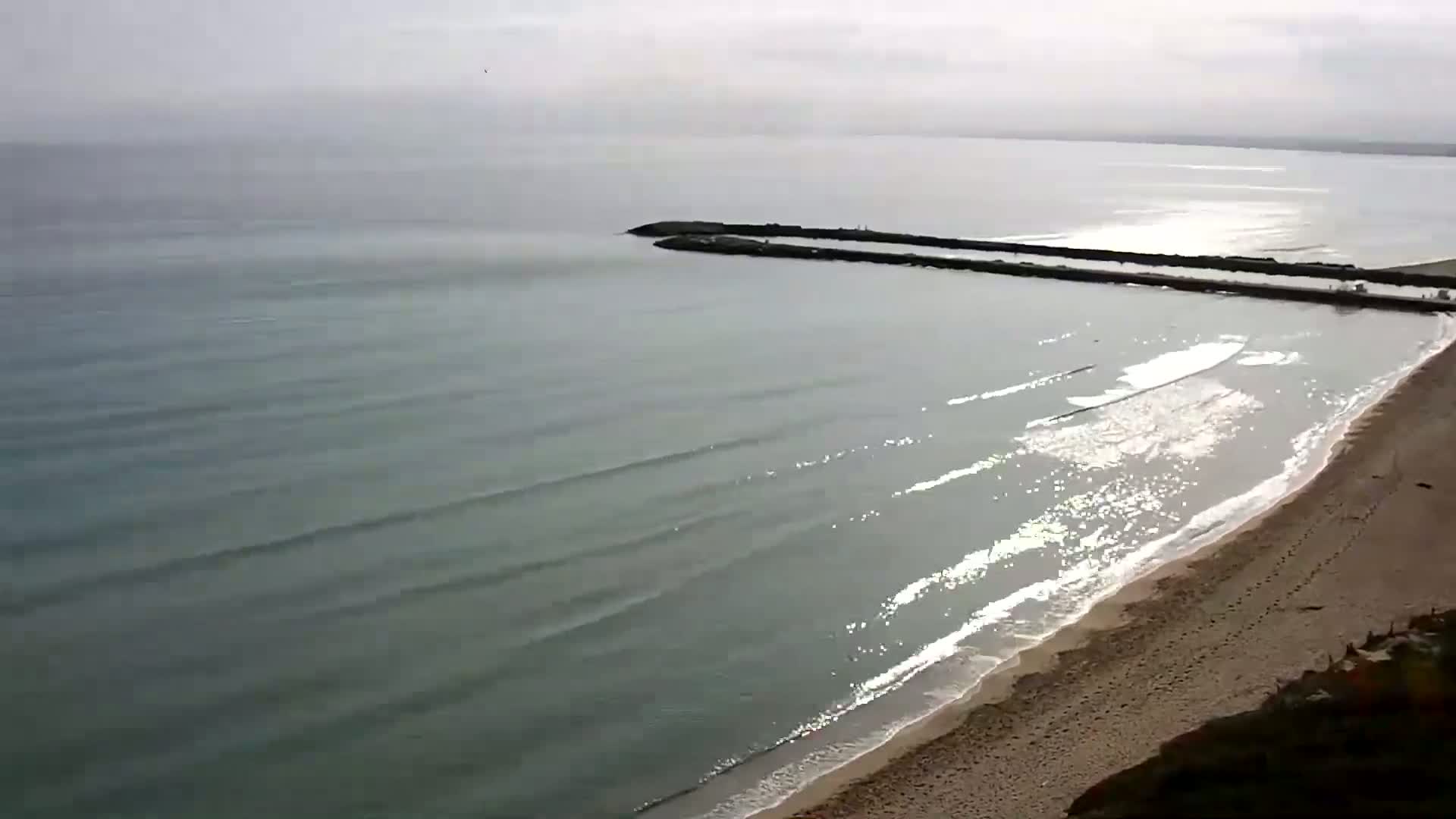 A sandy beach with gentle waves laps the shore under a cloudy sky, near a long, dark breakwater extending into a calm sea.