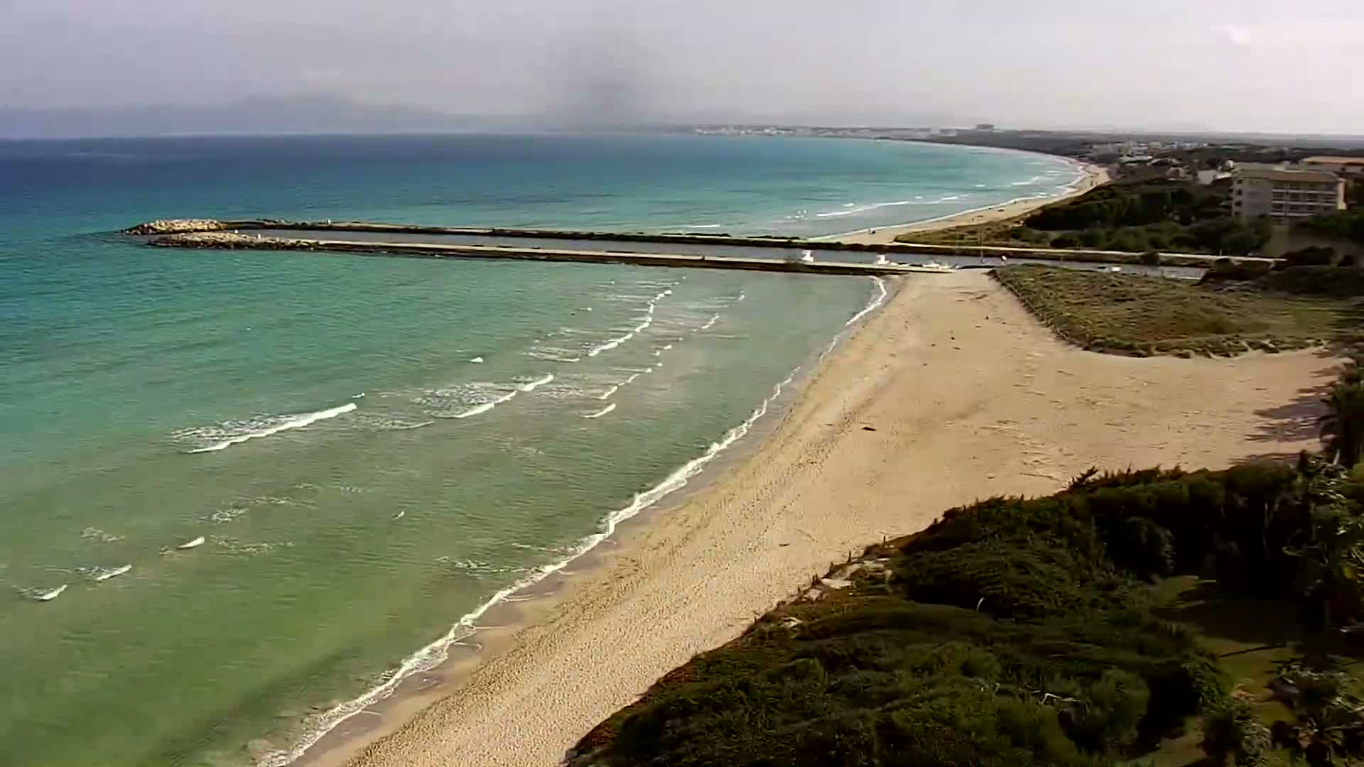 An aerial view shows a sandy beach meeting a calm, light-green ocean with small waves under a mostly cloudy sky.
