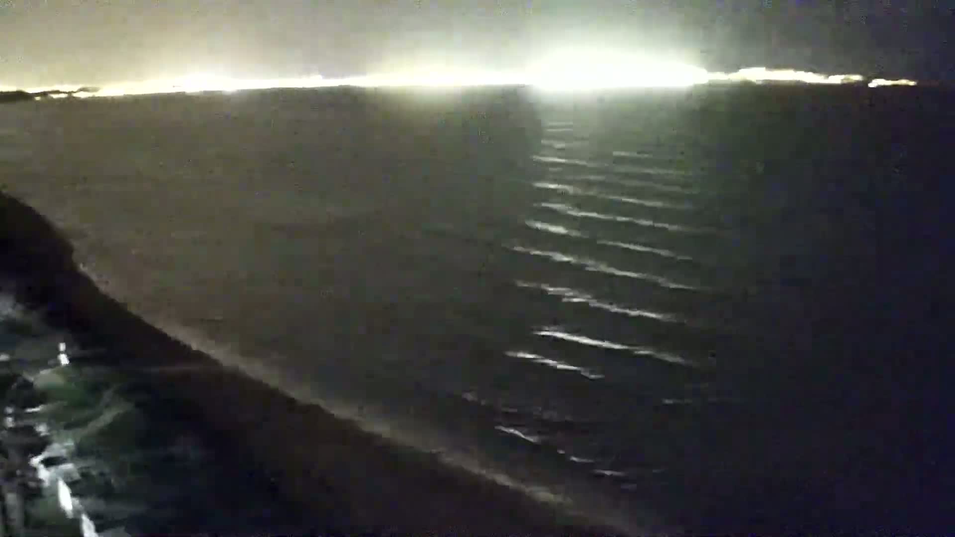 A nighttime view shows gently rolling waves on a dark sea under a dark sky, with a distant bright city glow on the horizon.