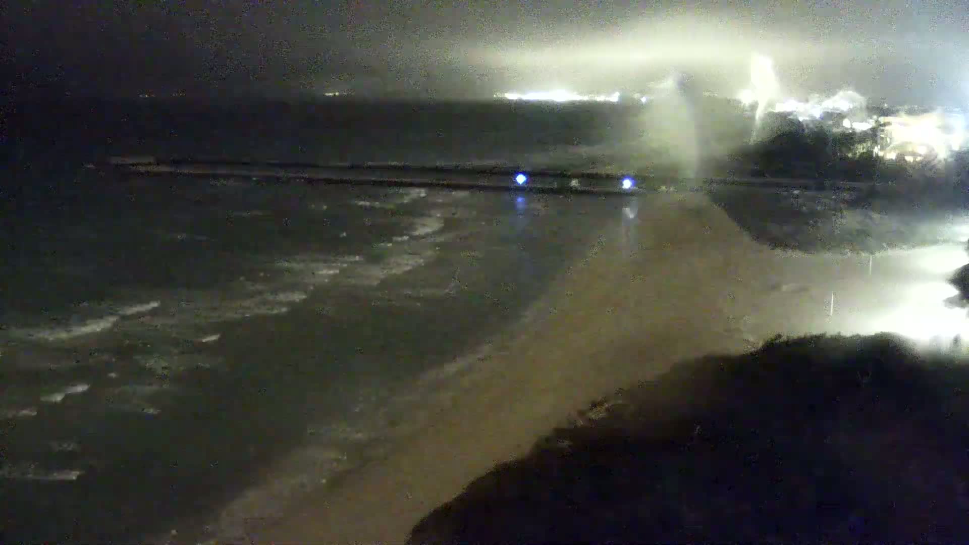 A nighttime view of a beach with dark, choppy water lapping at the shore under an overcast sky.