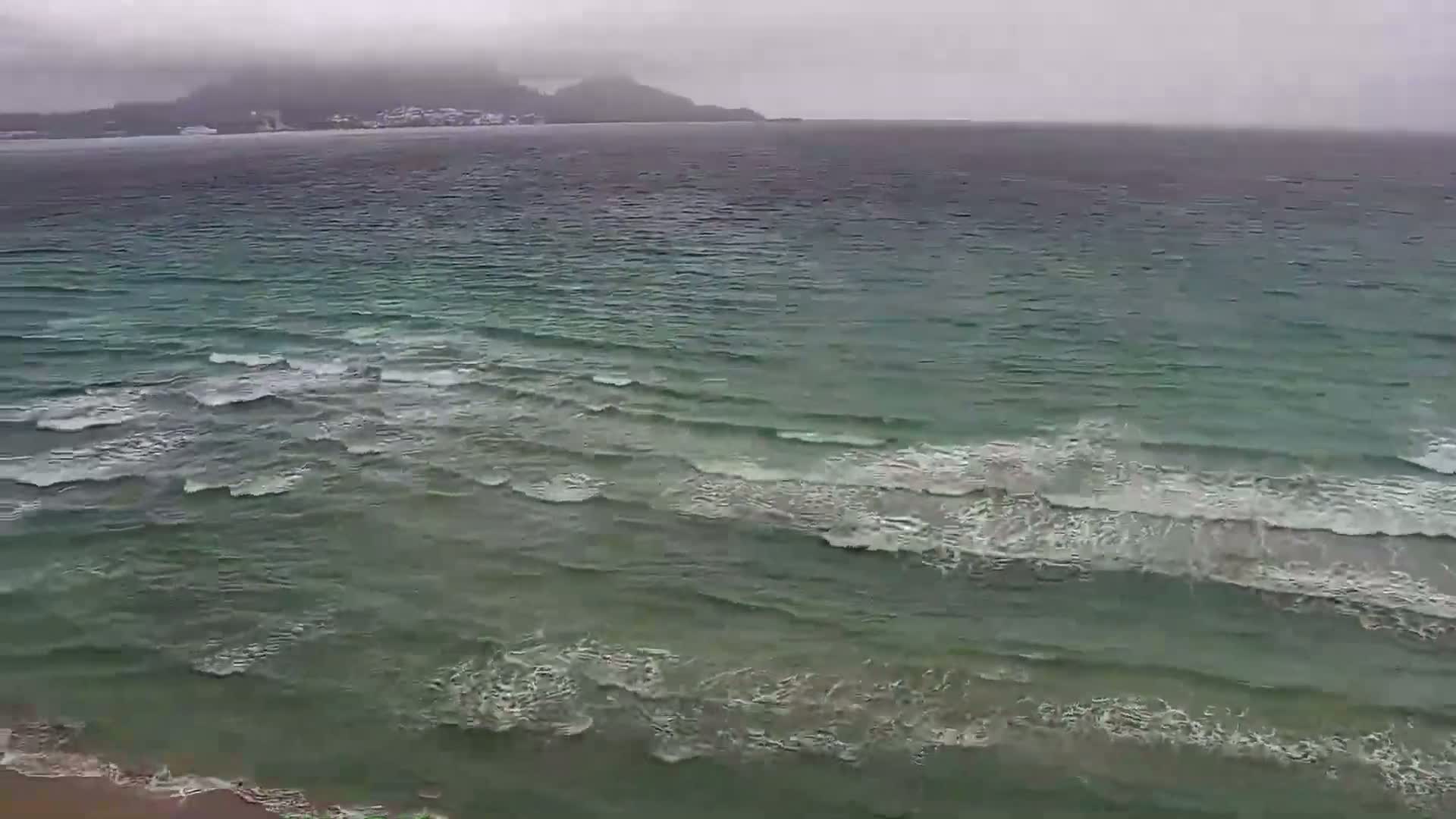 A hazy, overcast day shows a shoreline with moderate waves rolling into the beach, with a distant mountain range visible across the water.