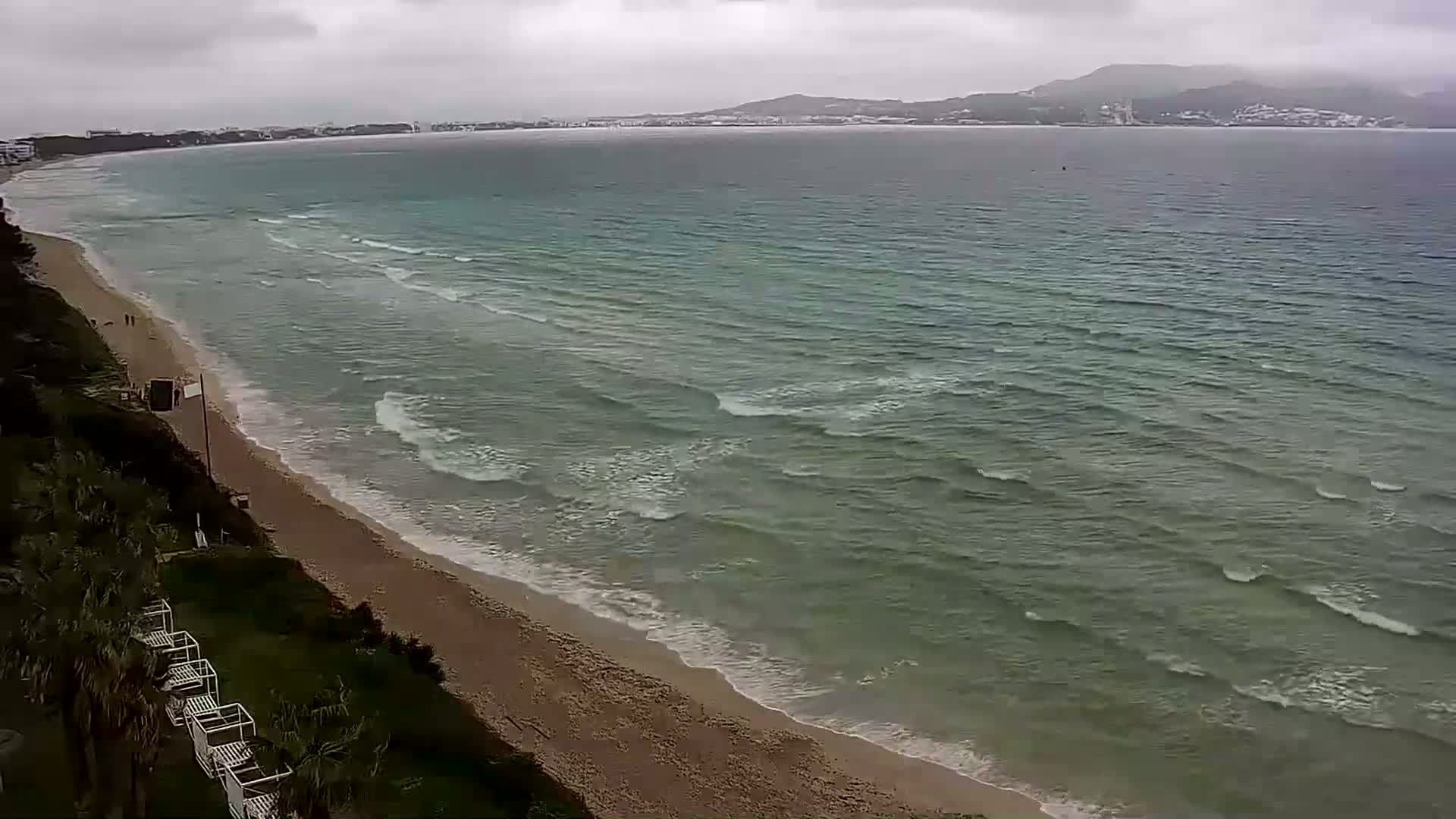 A sandy beach with gentle waves laps the shore under an overcast sky, with a distant coastline visible across the water.