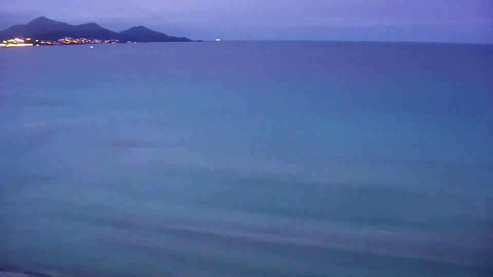 A calm, dark-blue ocean stretches out to a distant, dimly lit shoreline under a twilight sky.