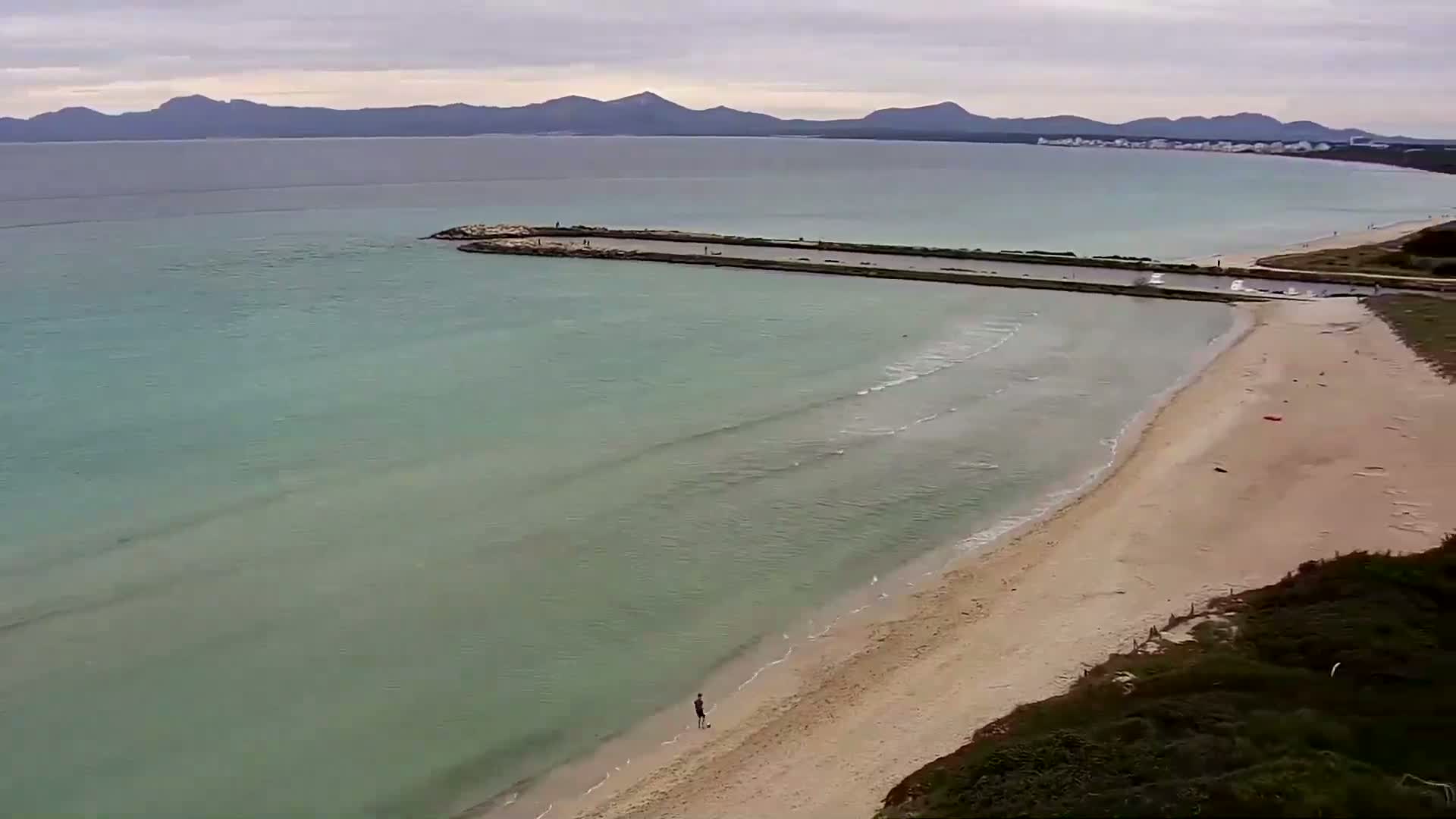 An aerial view shows a calm, light-green ocean lapping a sandy beach under an overcast sky, with mountains visible in the distance.