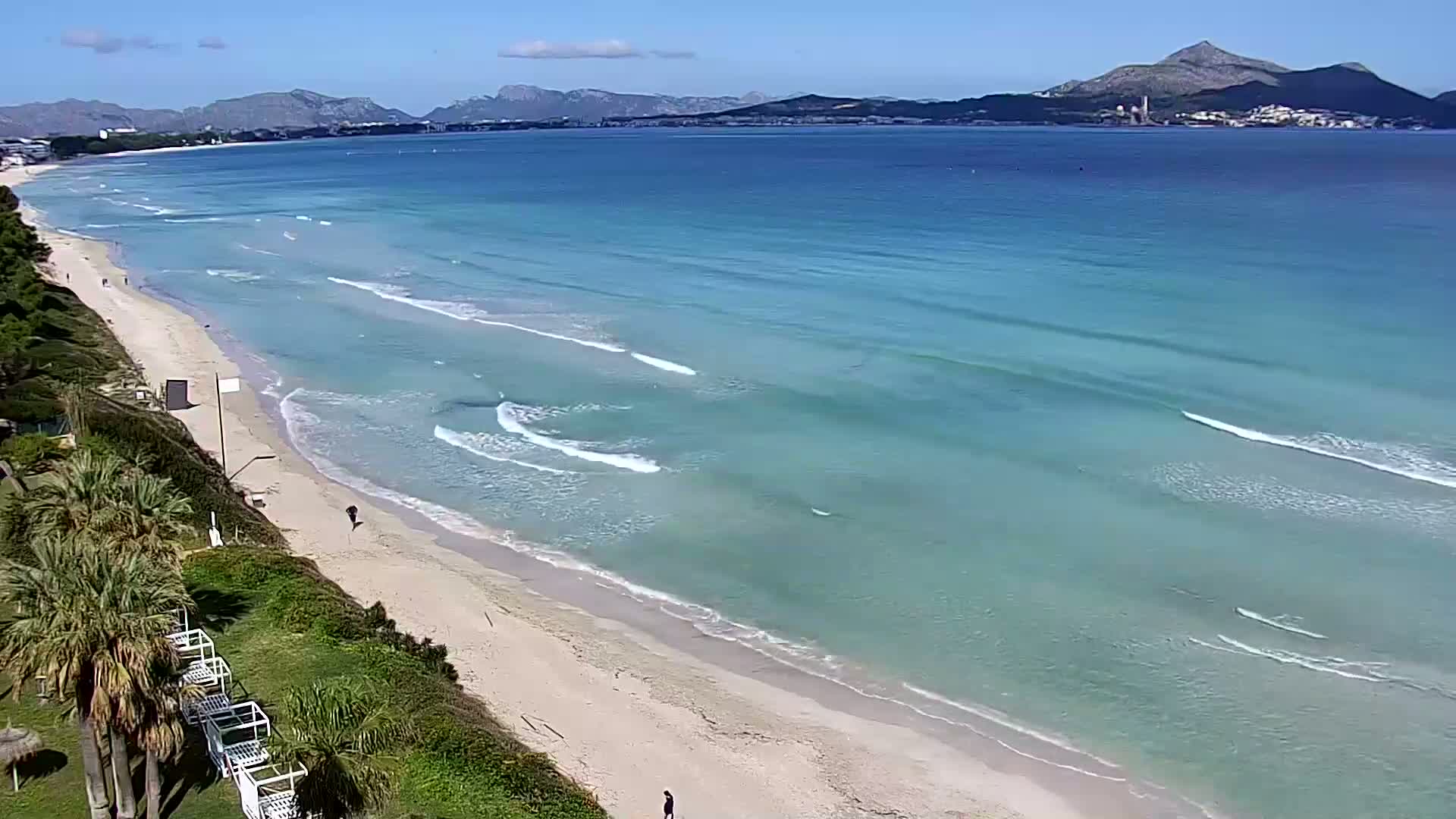A long, sandy beach with gently rolling waves stretches toward a calm sea under a clear, sunny sky, backed by distant mountains.