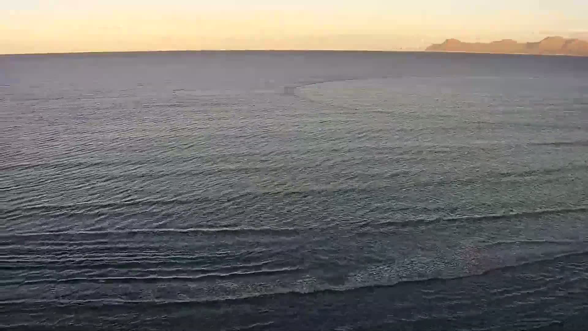 A calm, dark-blue ocean with small waves stretches to a distant, hazy shoreline under a pale orange-tinged sky.