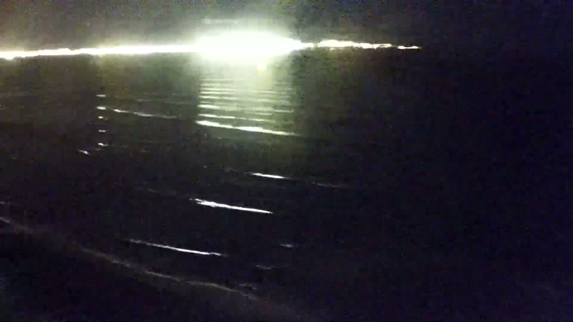 Dark, gently rippling water reflects a bright light source in the distance at night.