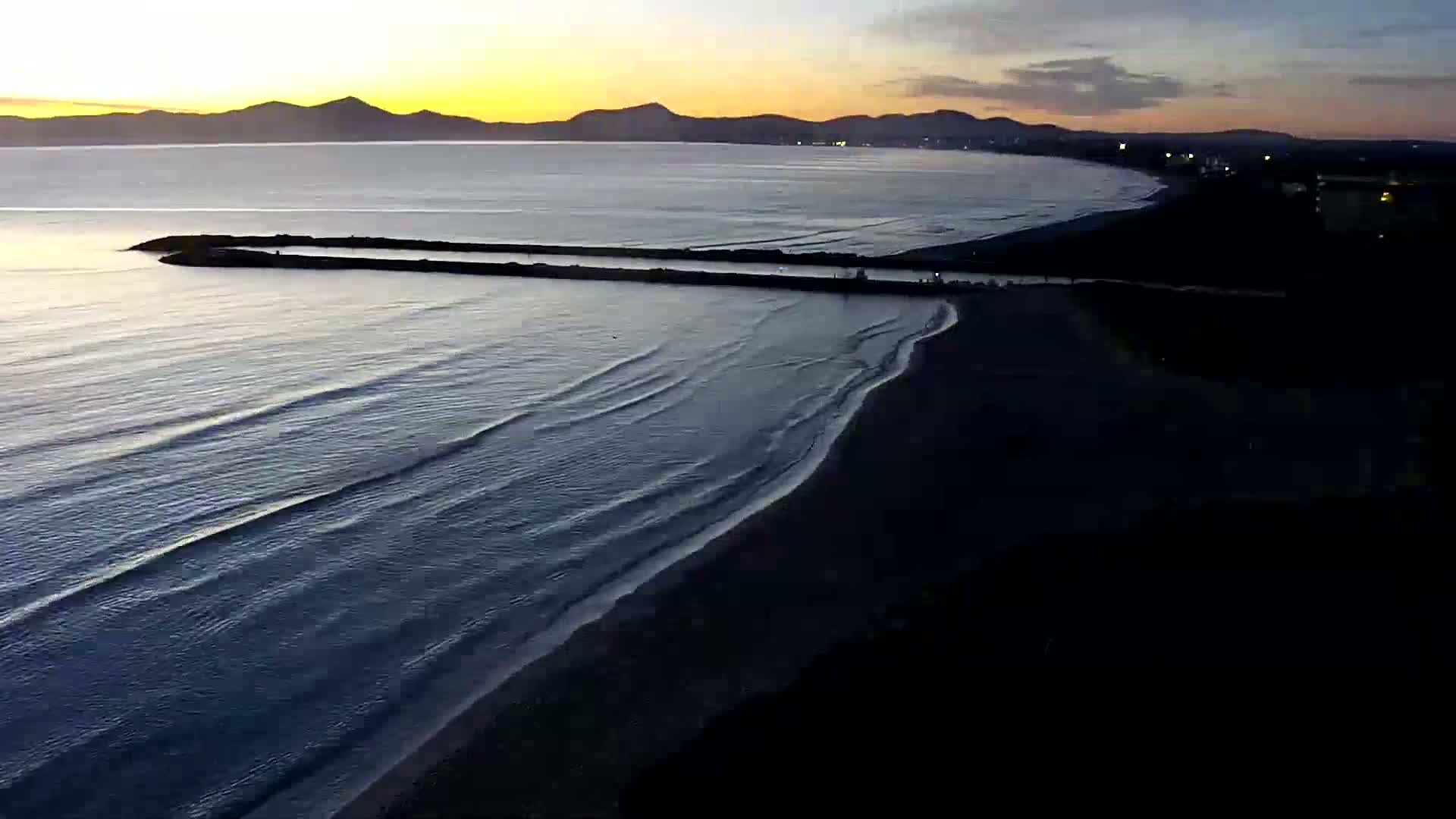 Gentle waves lap a dark beach at sunset, with a long jetty extending into a calm sea under a partly cloudy sky.