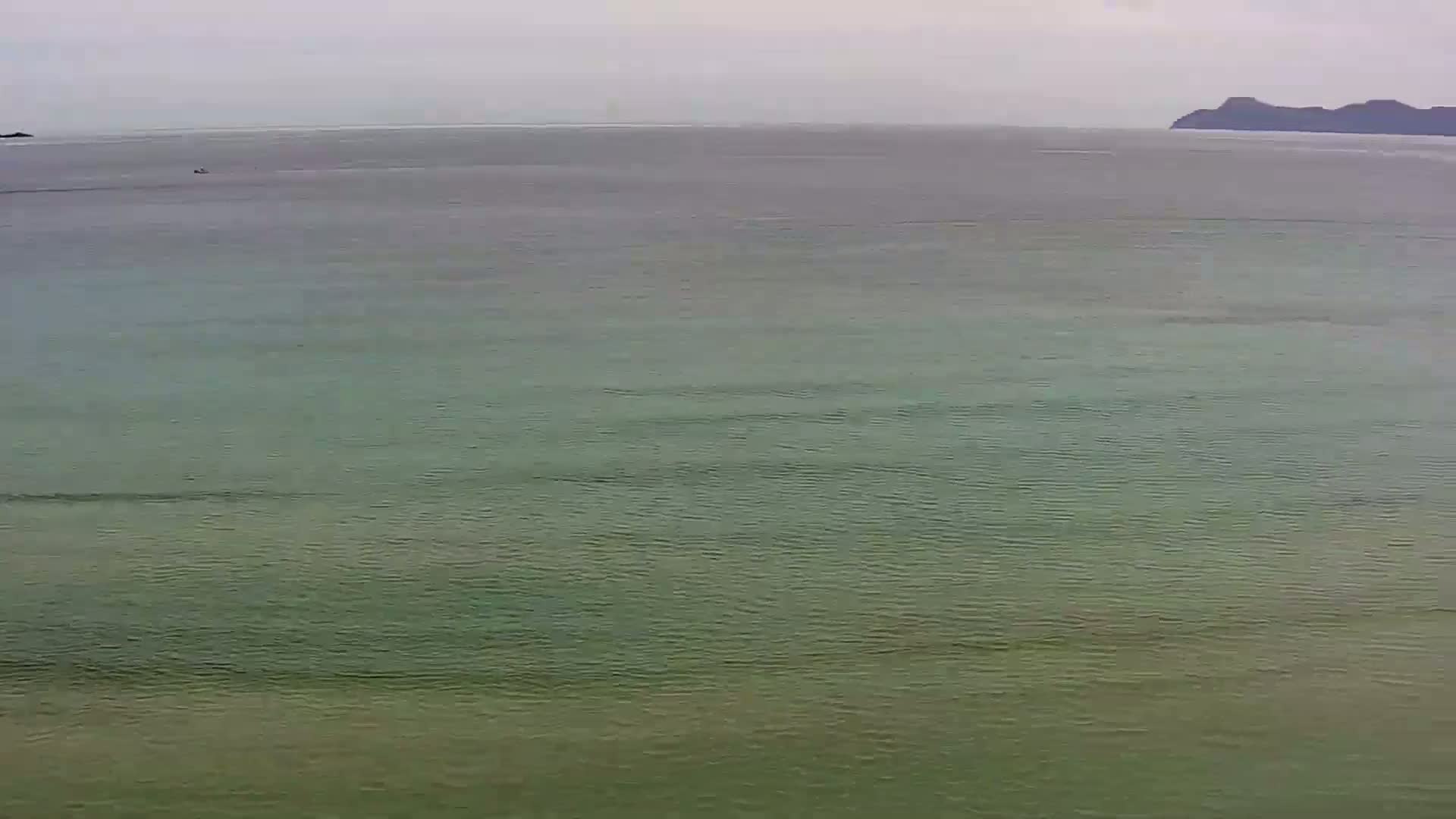 A calm, green-tinged ocean stretches to a distant, dark landmass under an overcast sky.