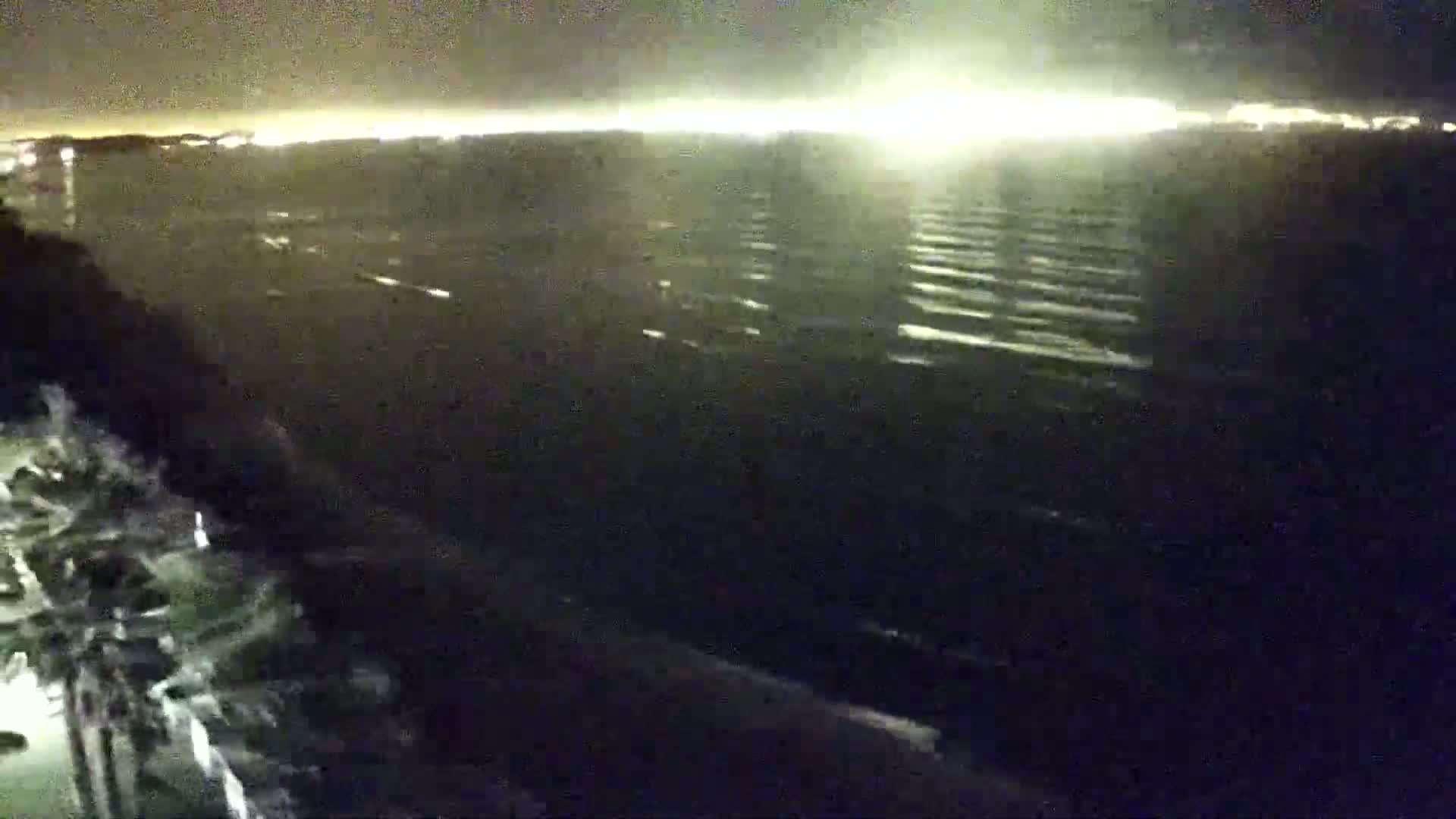 A nighttime view of a dark body of water with gentle waves, showing a distant shoreline illuminated by lights.