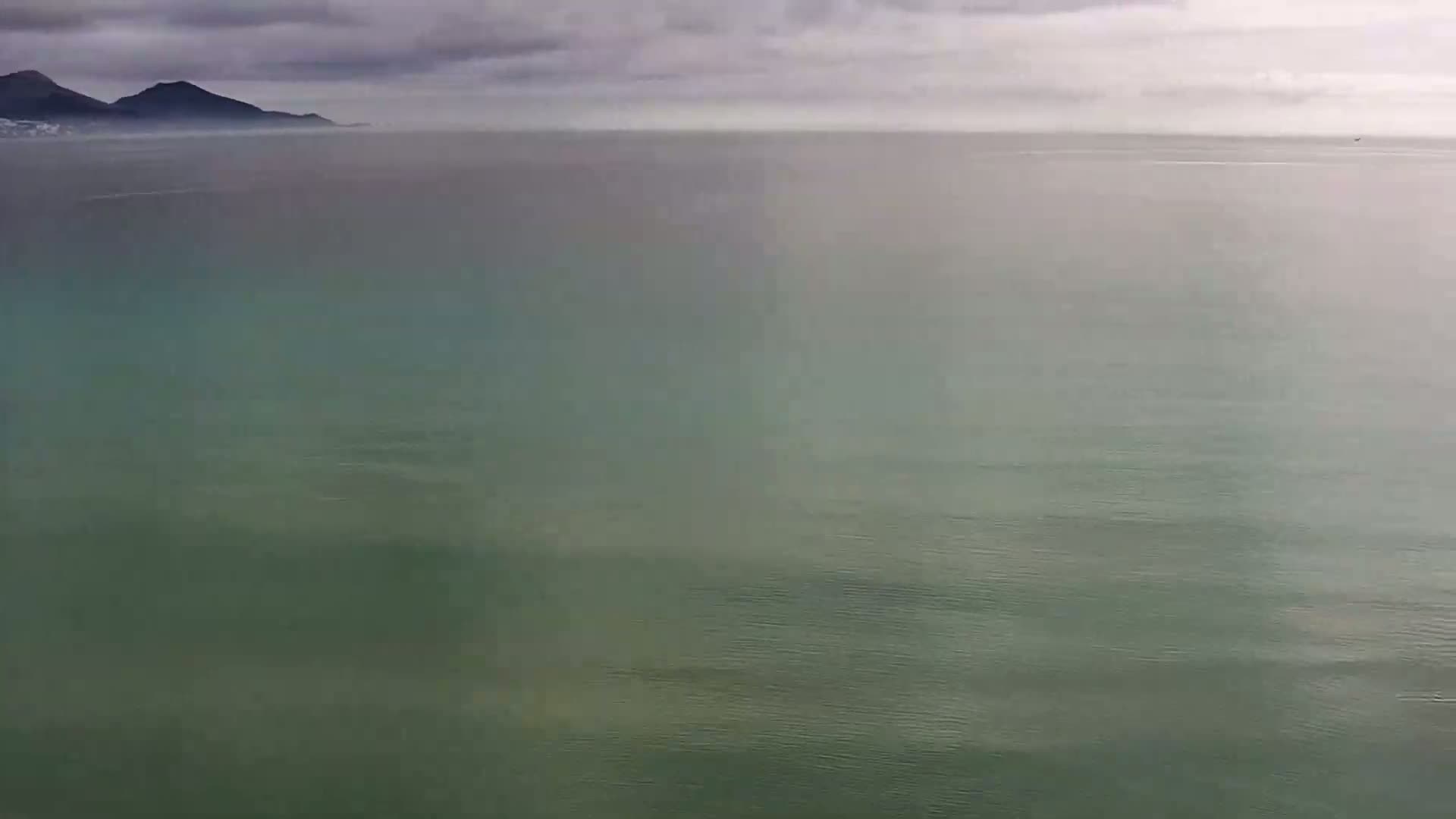 A calm, greenish-grey sea stretches to a hazy line of mountains under a cloudy sky.