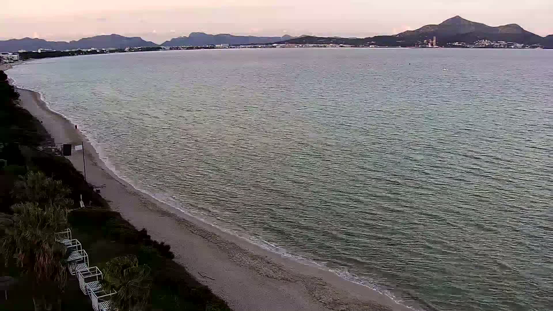 An aerial view shows a sandy beach meeting calm, gray-green water under an overcast sky, with distant mountains visible across the bay.