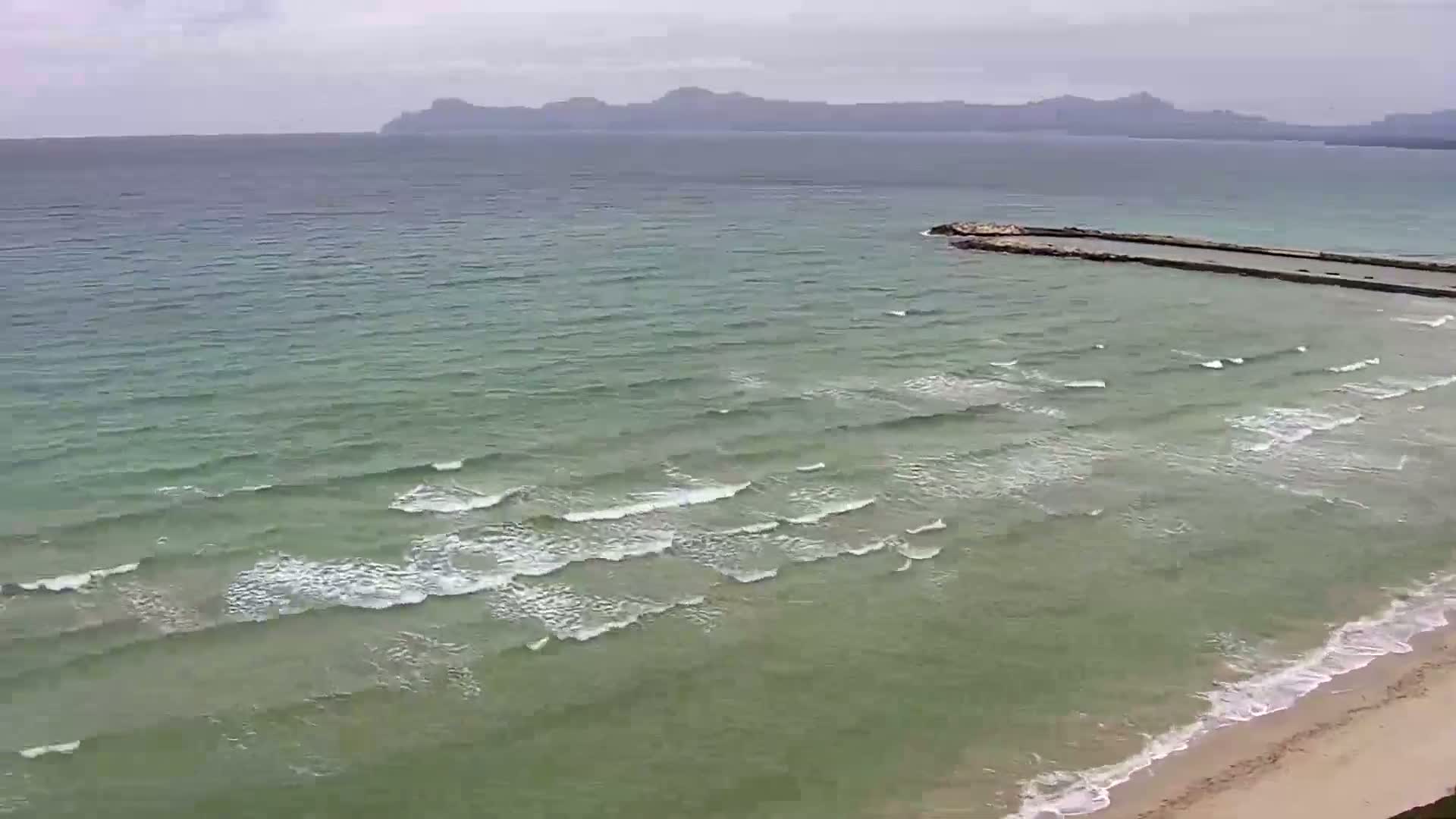 A long, low-lying breakwater extends into a sea with small waves under an overcast sky, near a sandy beach and distant mountains.