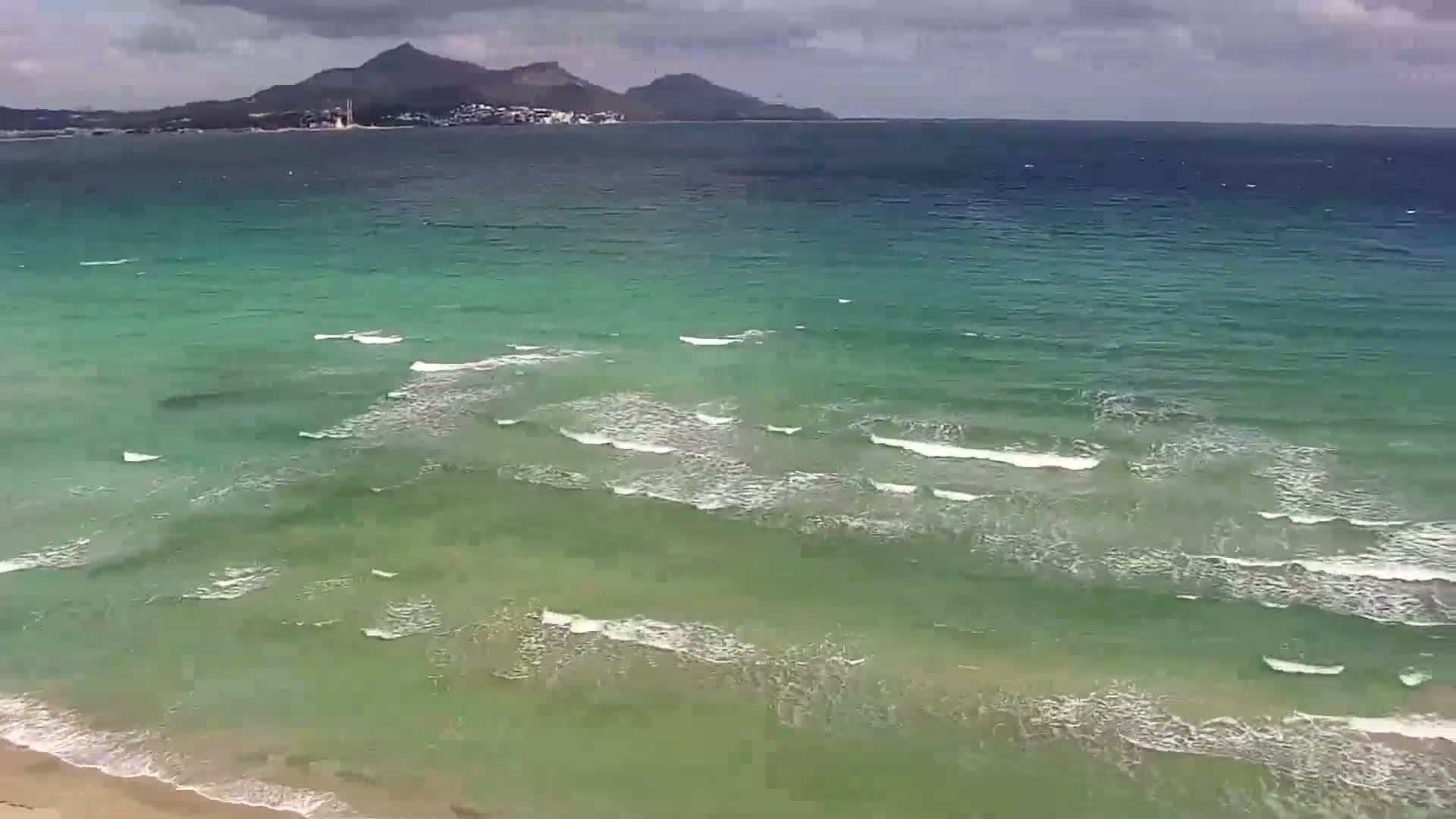 A partly cloudy day shows a wide expanse of teal-green ocean water with small waves lapping a sandy beach, and distant mountains visible on the horizon.