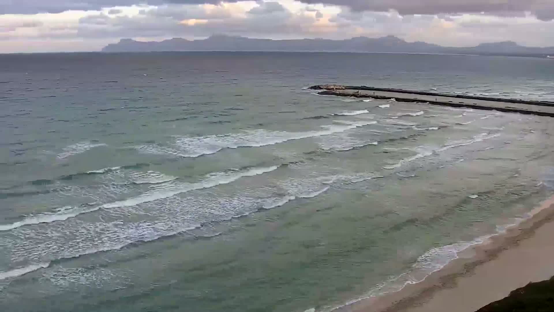 A beach with gently rolling waves under a cloudy sky, a low-lying jetty extends into the sea with distant mountains visible on the horizon.