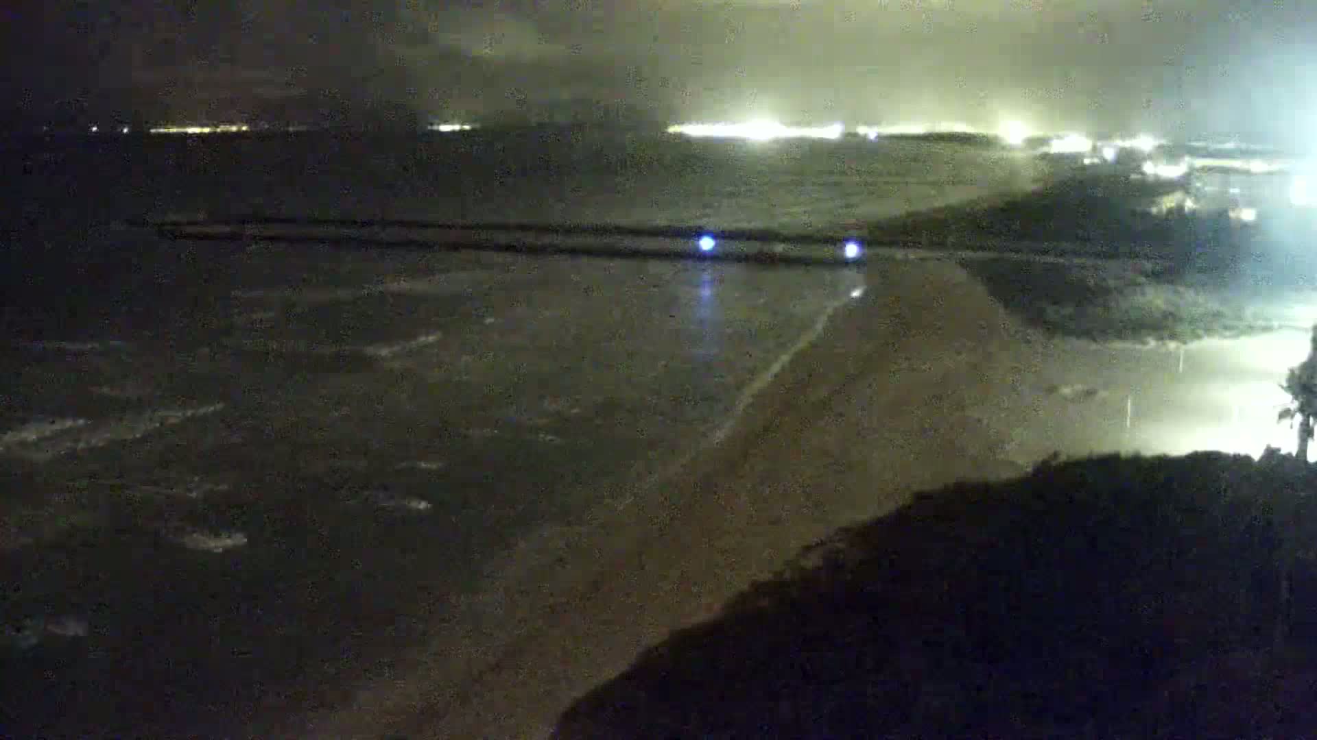 A nighttime view of a dark, wavy ocean meeting a sandy shore under a cloudy sky, with lights visible in the distance.