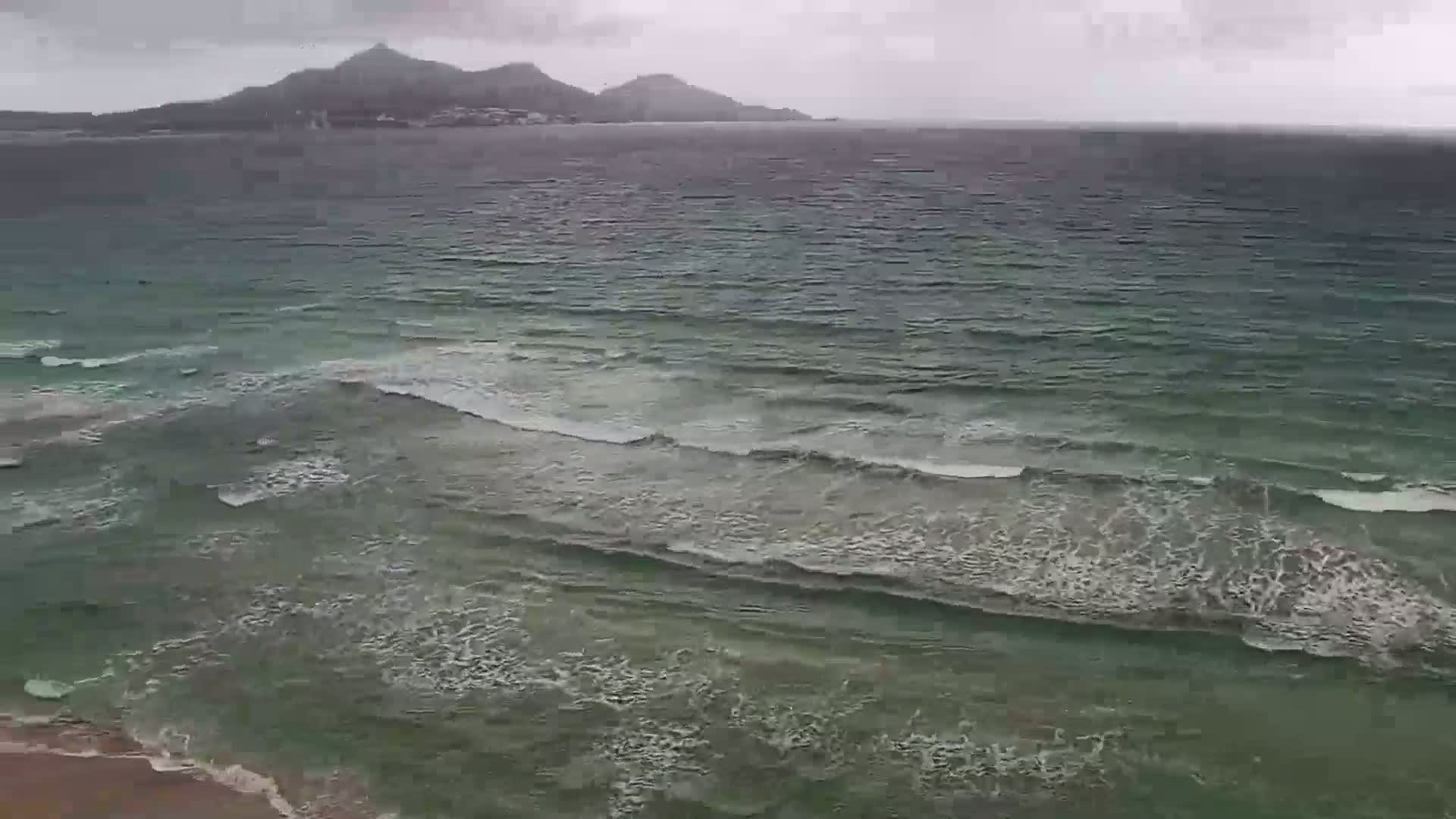 Gentle waves roll onto a sandy beach under an overcast sky, with hills visible in the distance.