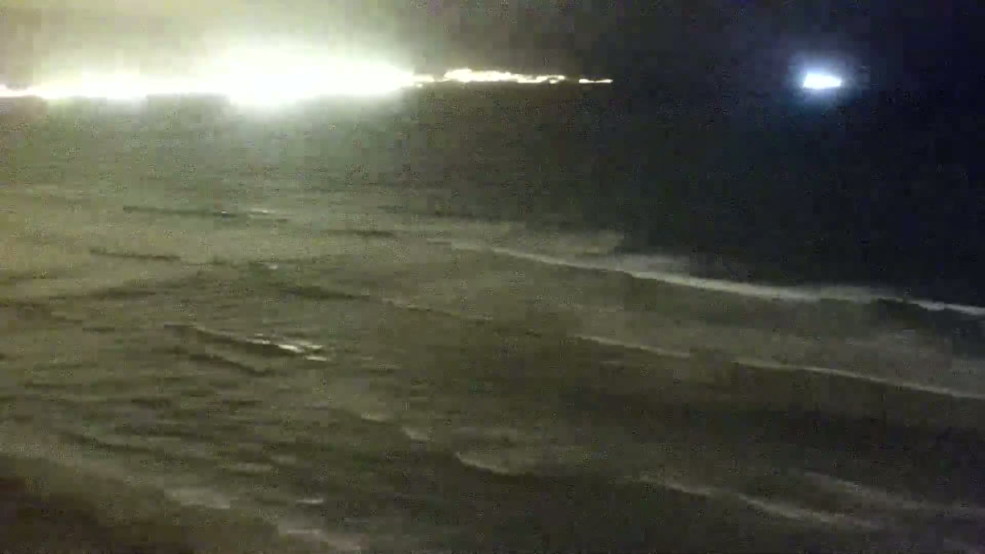 A nighttime view of a dark ocean with waves under a mostly cloudy sky, illuminated by distant lights and a bright moon.