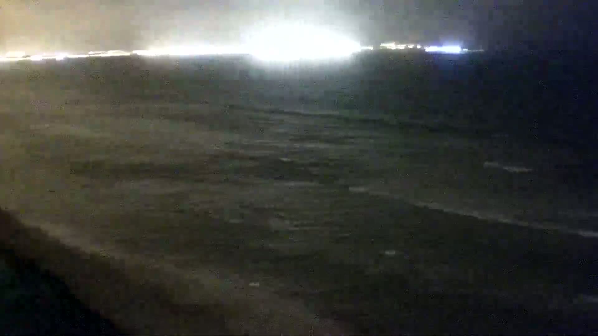 A nighttime, low-light view of a dark ocean with small waves, illuminated by distant city lights on the horizon.