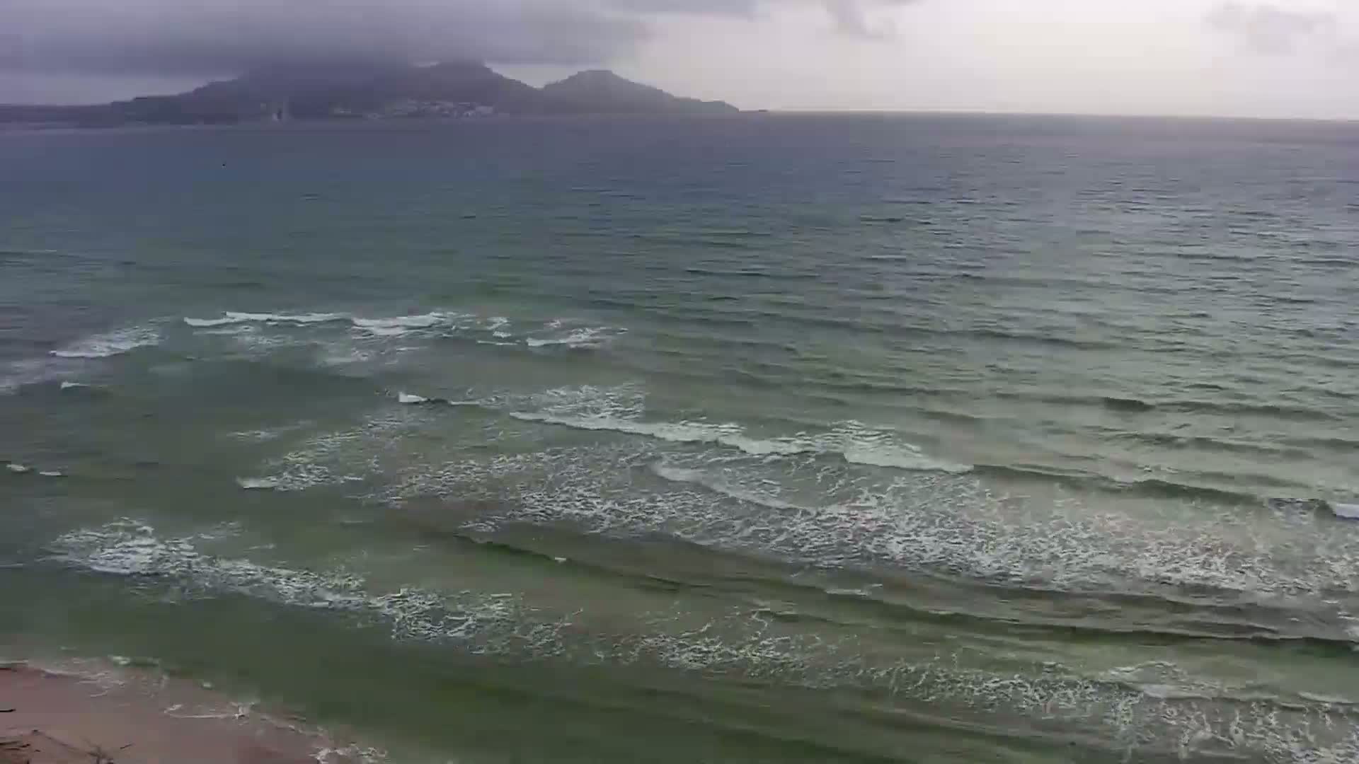 A beach with moderate waves rolls onto the shore under an overcast sky, with a distant mountain visible on the horizon.