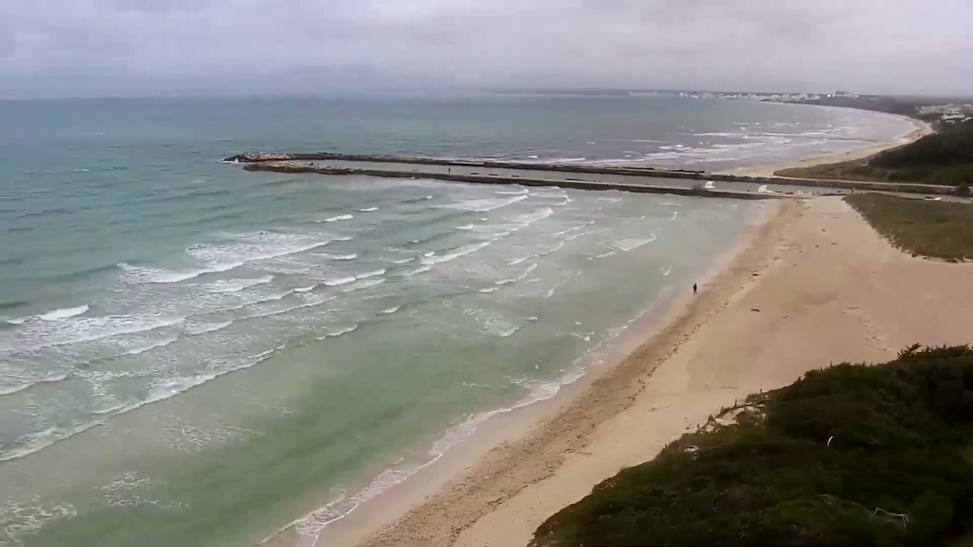 An expansive sandy beach meets a calm, teal ocean with small waves under an overcast sky.