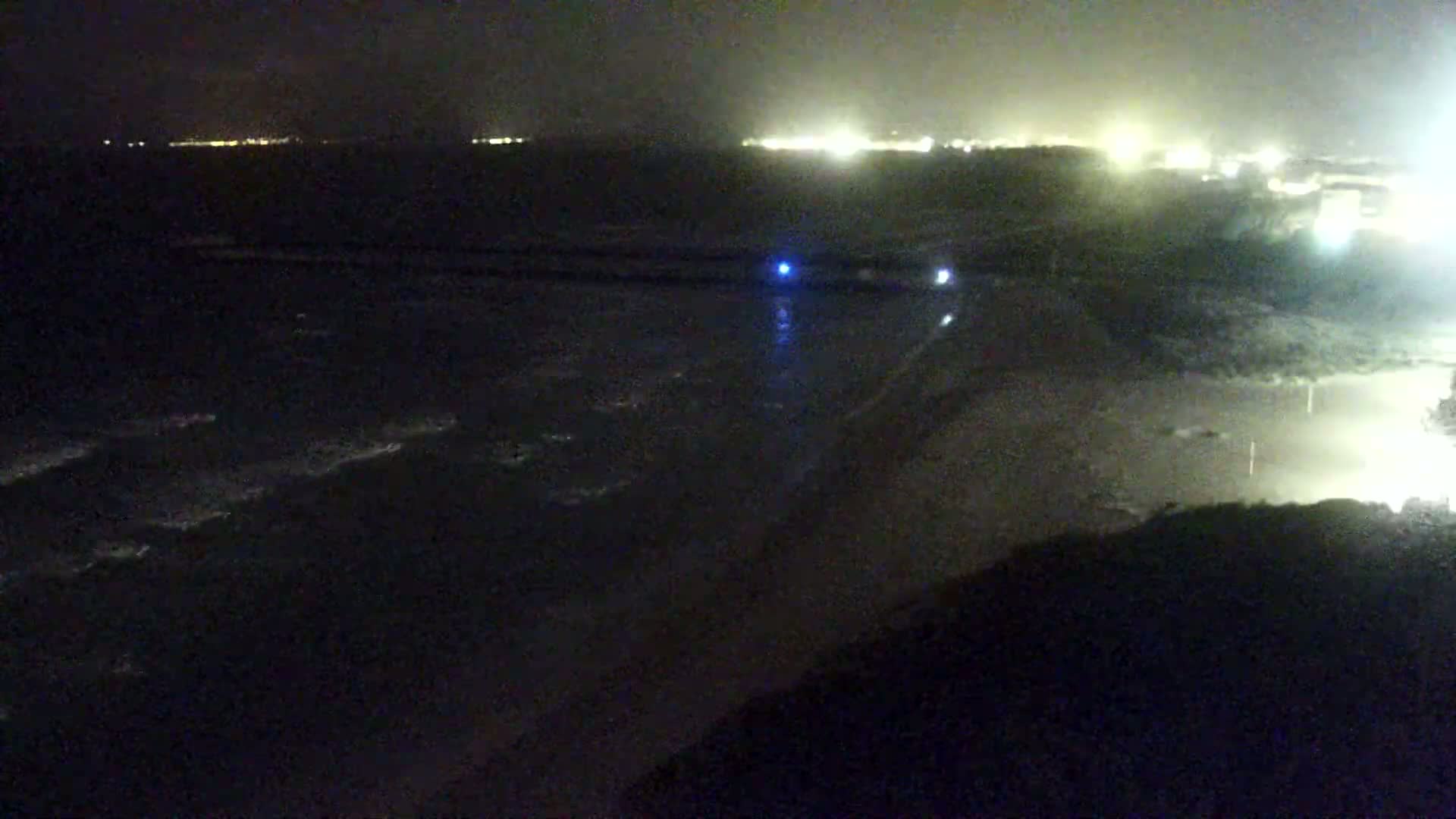 A dark, nighttime beach scene shows gently lapping waves on the shore with distant lights visible across the water.
