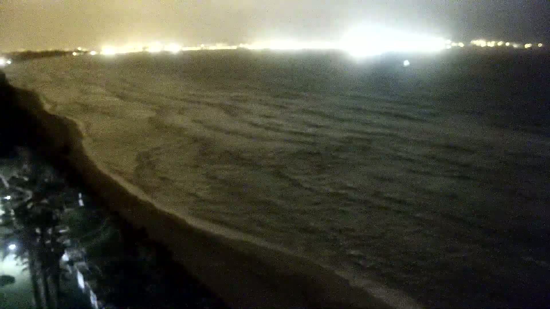 A nighttime view of a dark ocean with small waves lapping a sandy shore, illuminated by distant city lights.