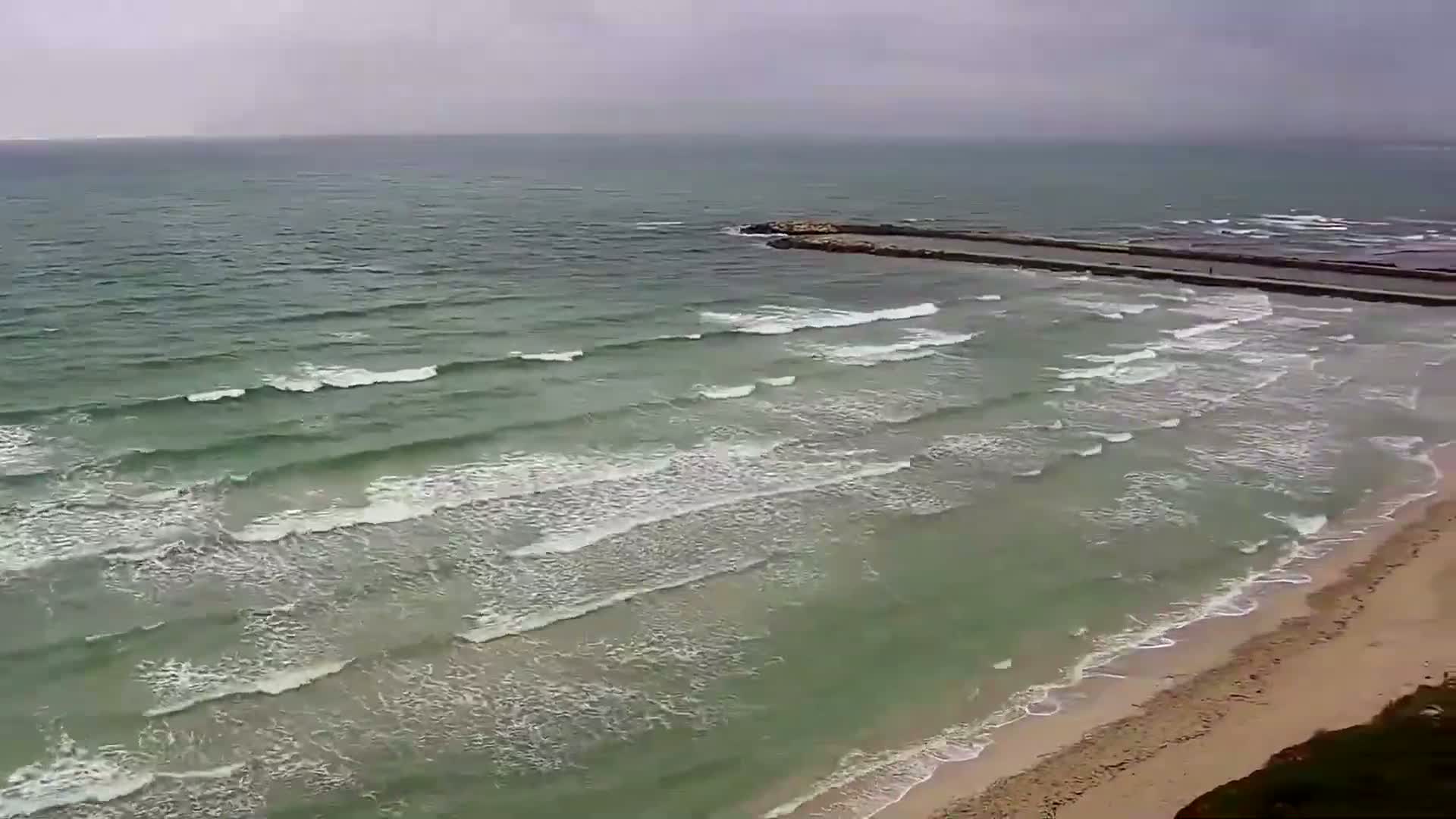 A sandy beach meets a greenish-grey sea with moderate waves under an overcast sky.
