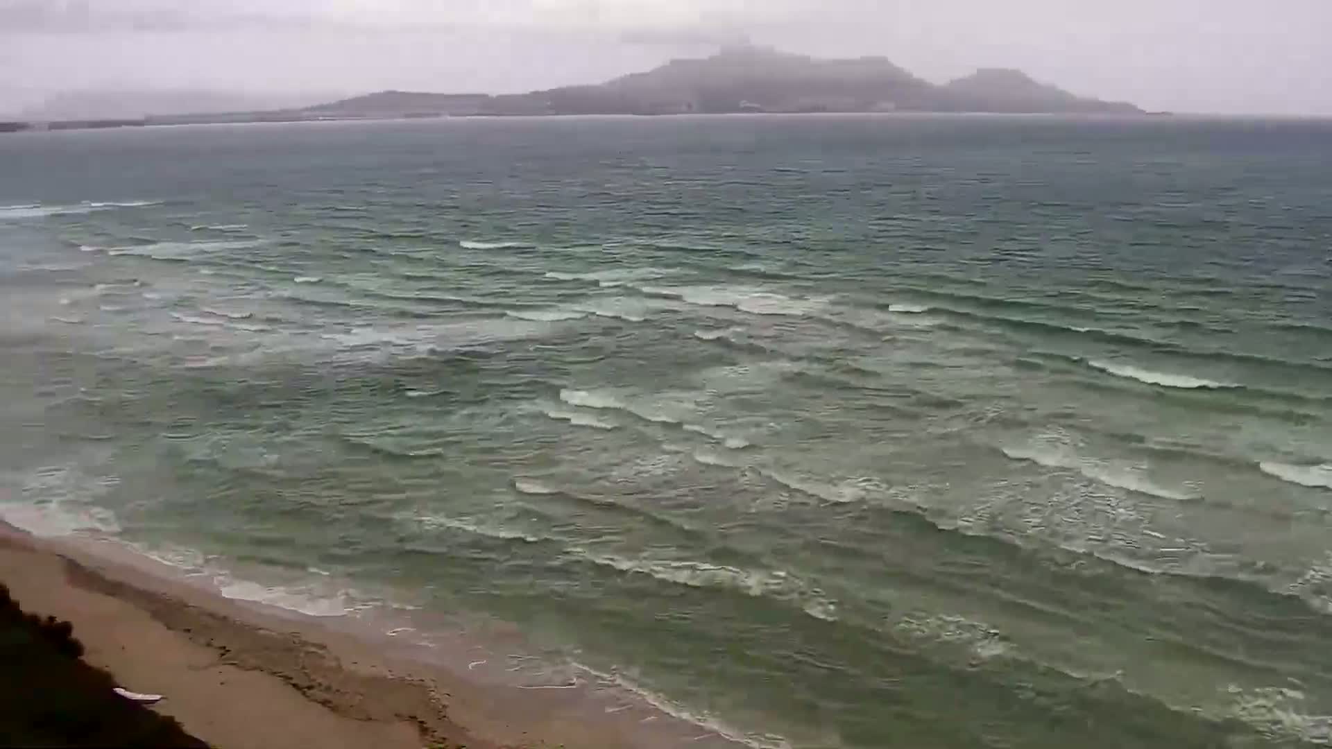 A sandy beach meets a sea with moderate waves under an overcast sky, with a distant mountain range visible on the horizon.